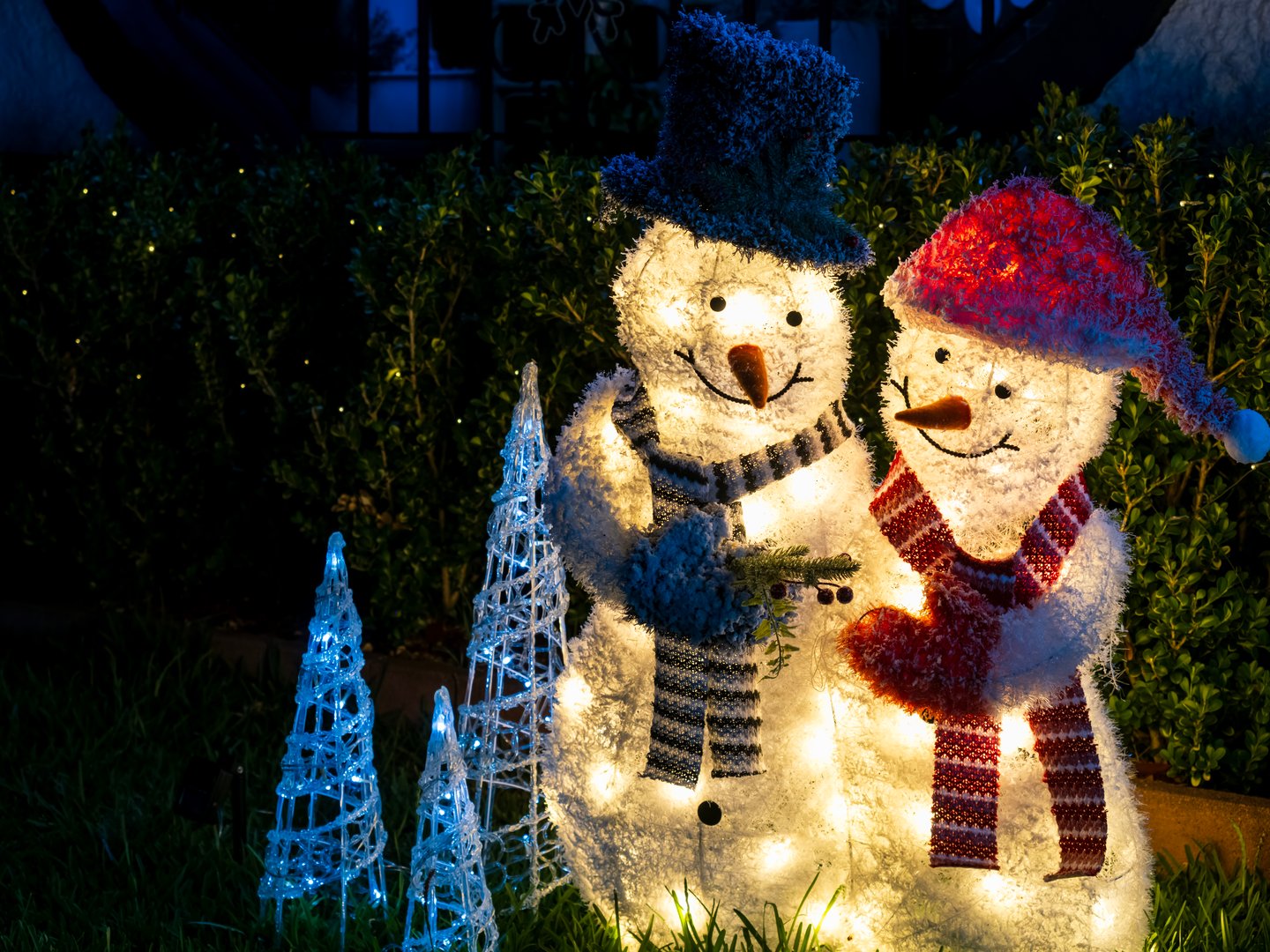 Two side-by-side effigies of illuminated Christmas Snowmen with festive decorative lights in front of a suburban house. View from a public pedestrian pathway.