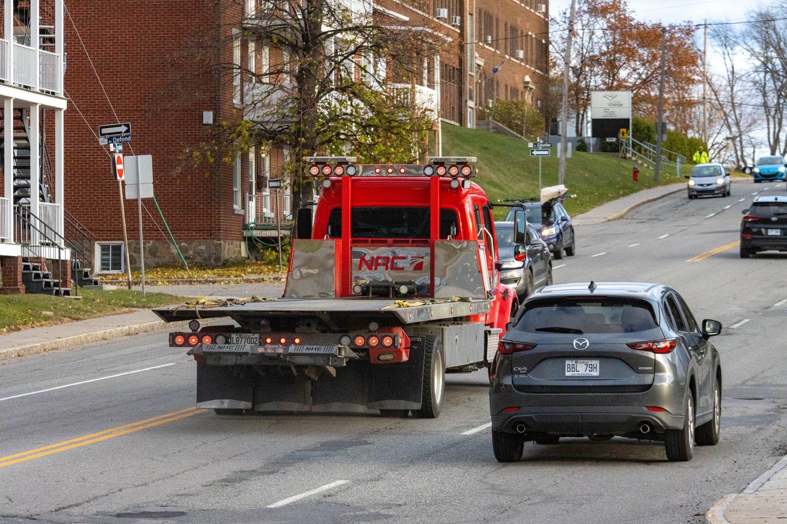 Canada, Quebec, 08 November 2025 : Red tow truck driving behind Mazda on city street afternoon traffic