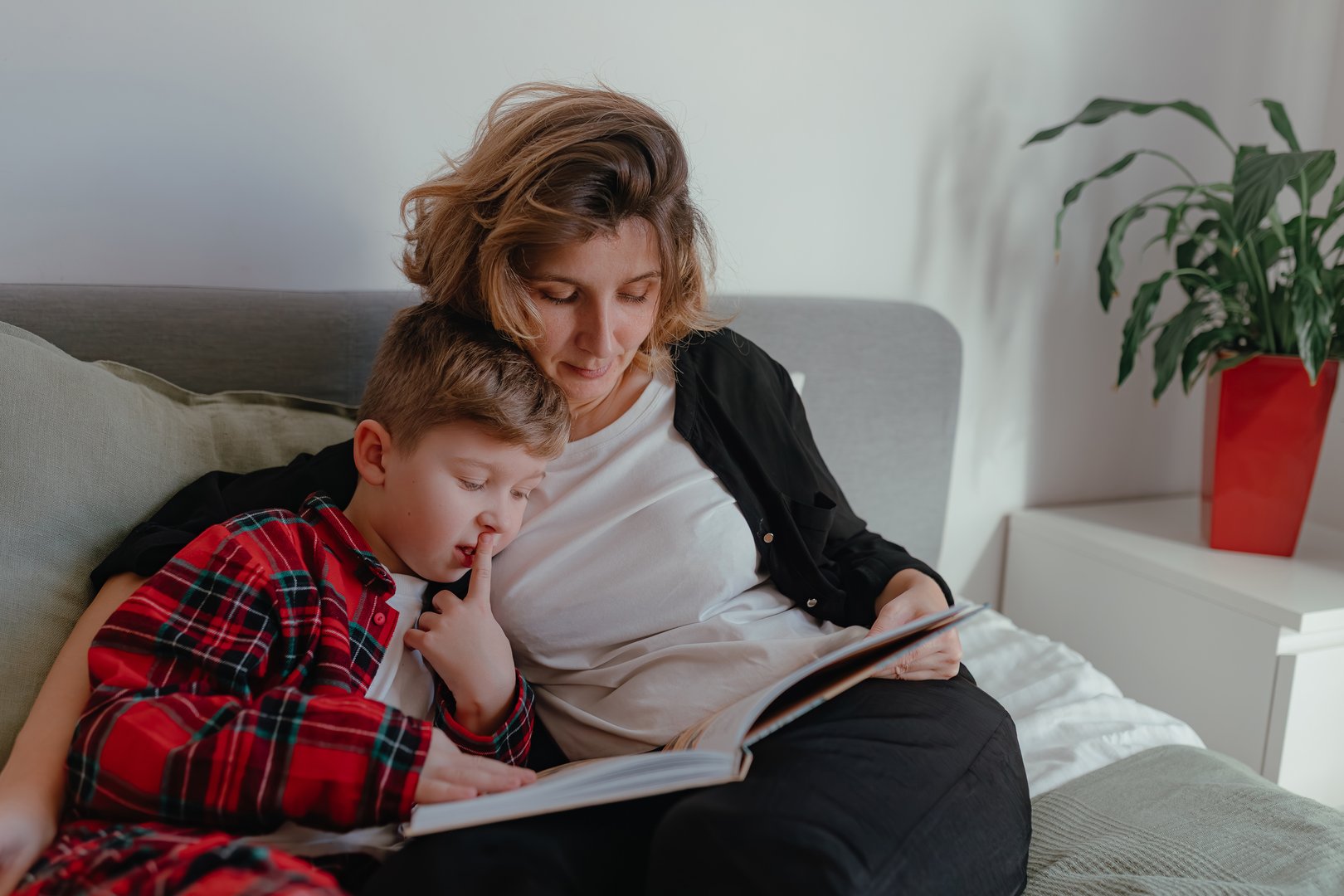 Mother supporting child while both focus over open storybook during soft daylight inside bright bedroom with gentle tones, pillows, spacious layout, peaceful shared reading moment.