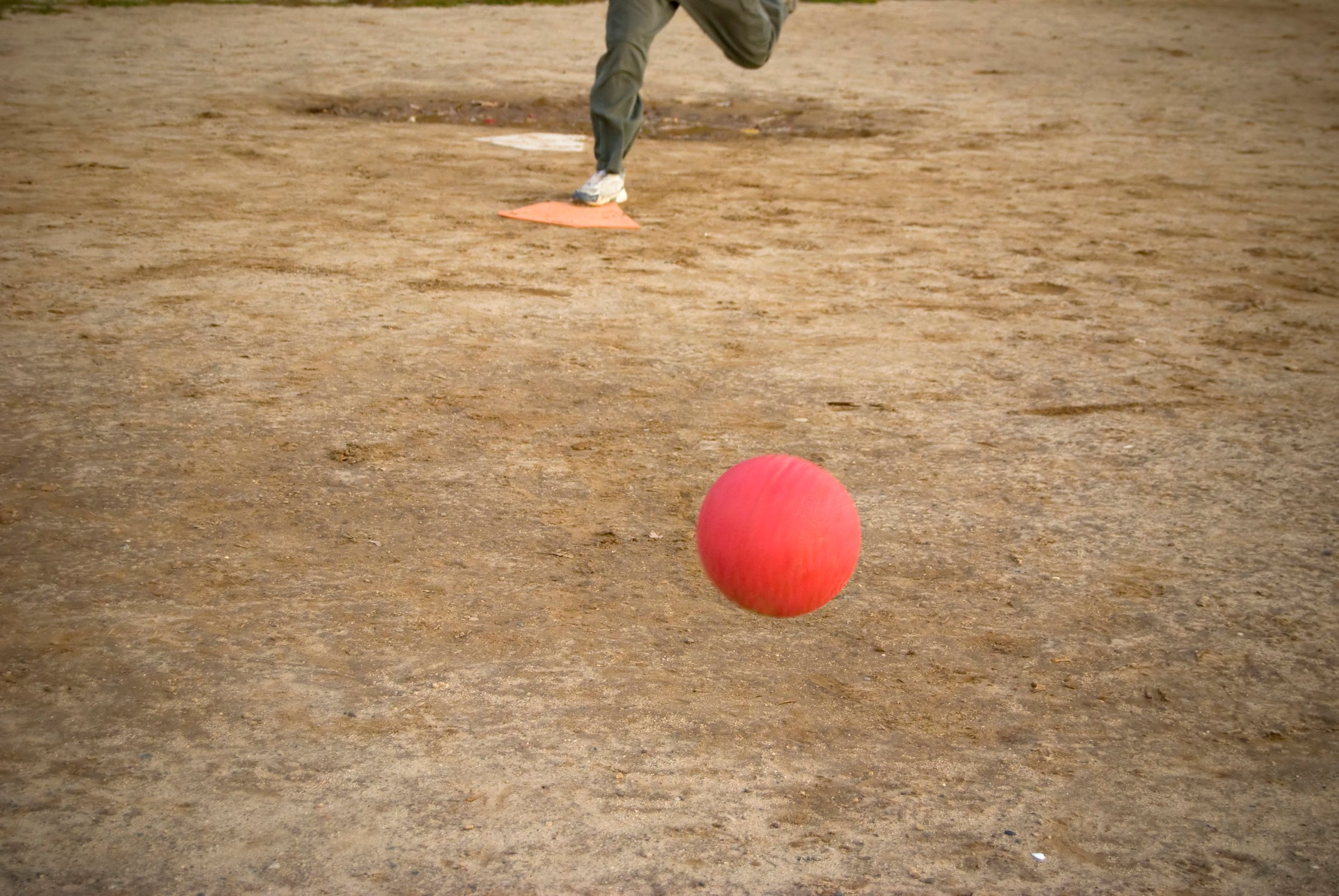 Person playing kickball on dirt field with red ball in foreground and orange base visible
