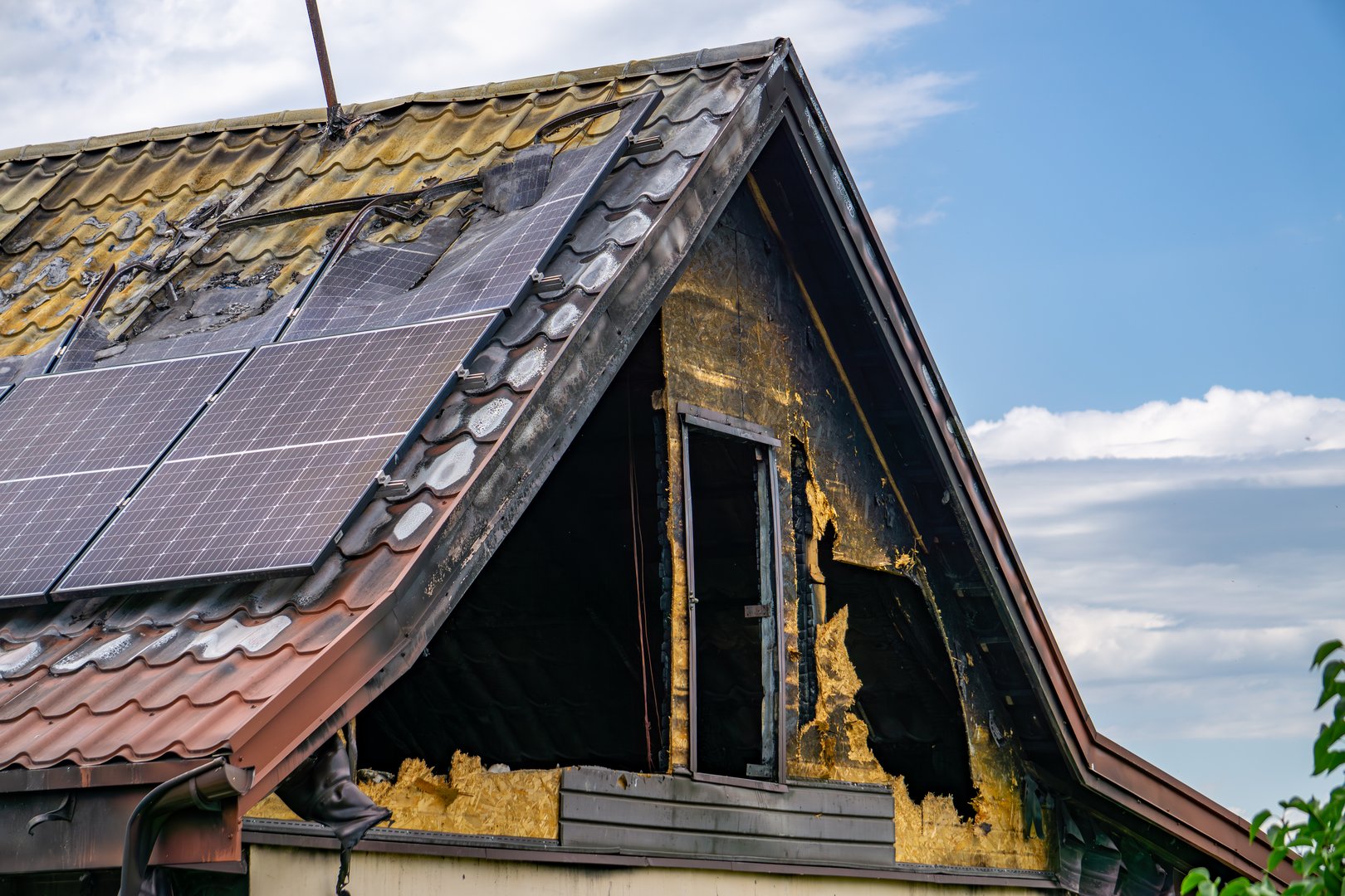 Burnt roof of a house with solar panels, showing fire damage on the attic and charred facade under a partly cloudy sky.