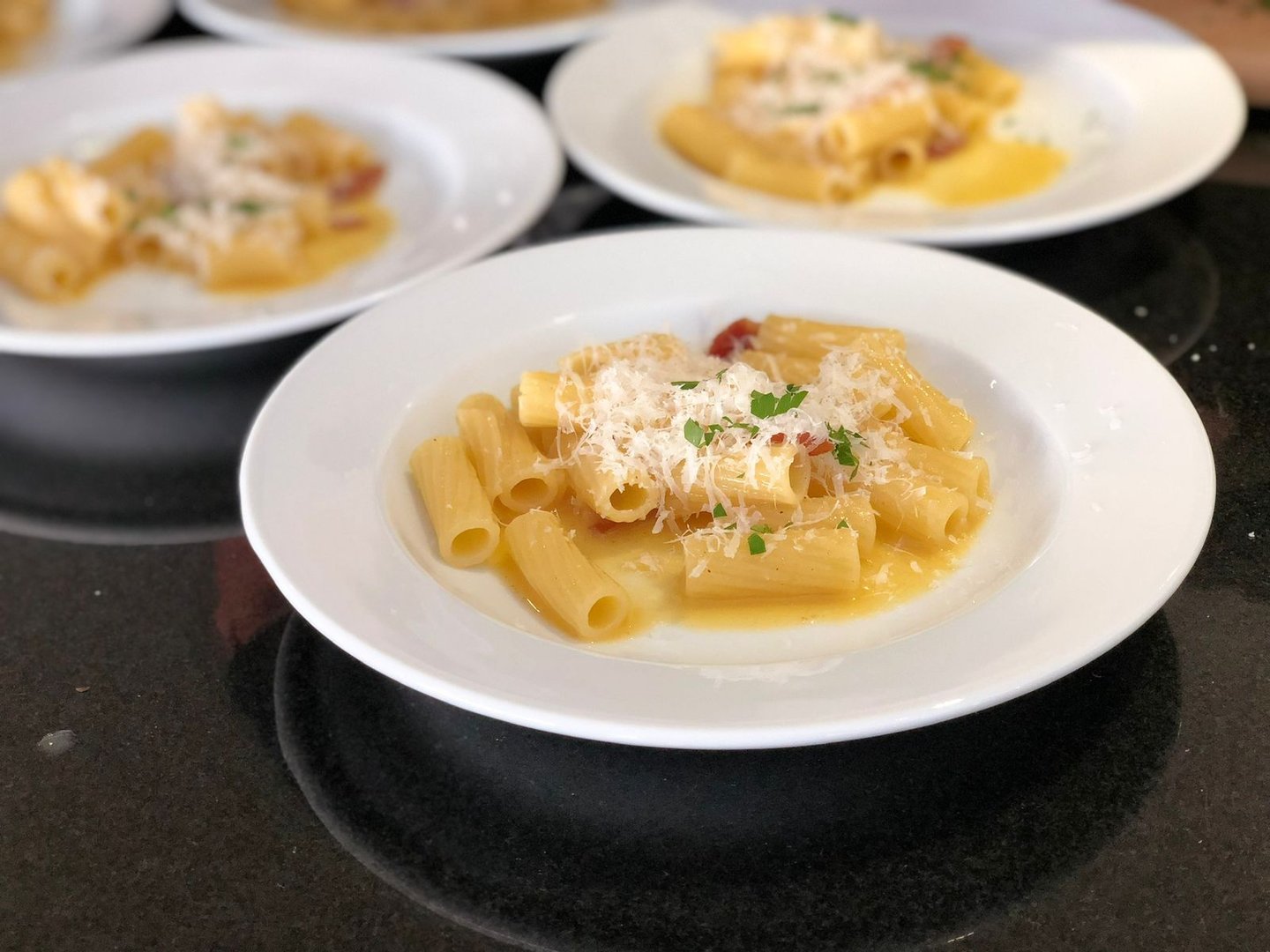 Plates of rigatoni pasta topped with grated cheese and herbs on a glossy black surface.
