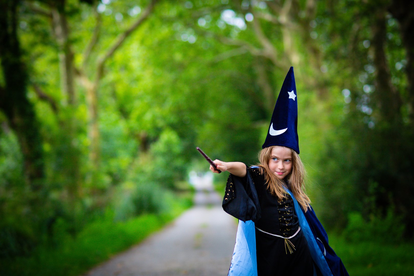 Caucasian blonde child girl in wizard blue costume with cape and hat, casting a spell with magic wand in outdoor forest background.