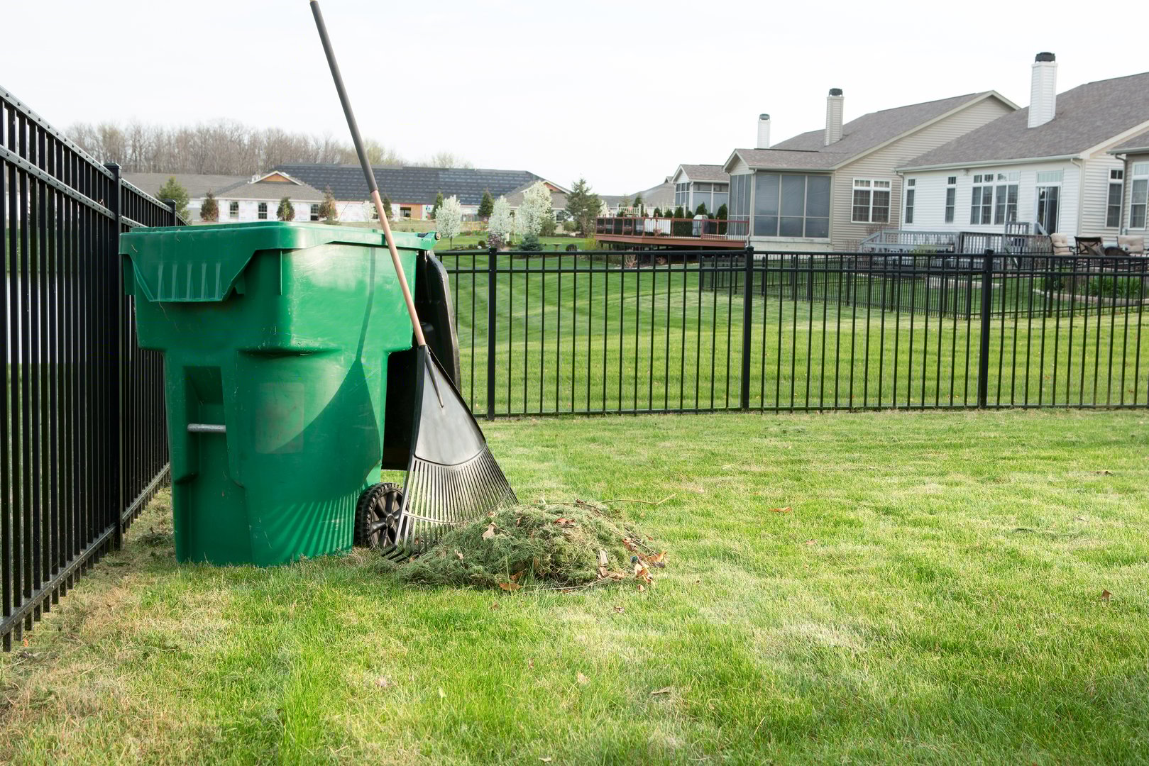 Raking lawn clippings on a neat upmarket suburban housing estate with a heap of grass cutting alongside a rake leaning on a green plastic bin for composting organic waste