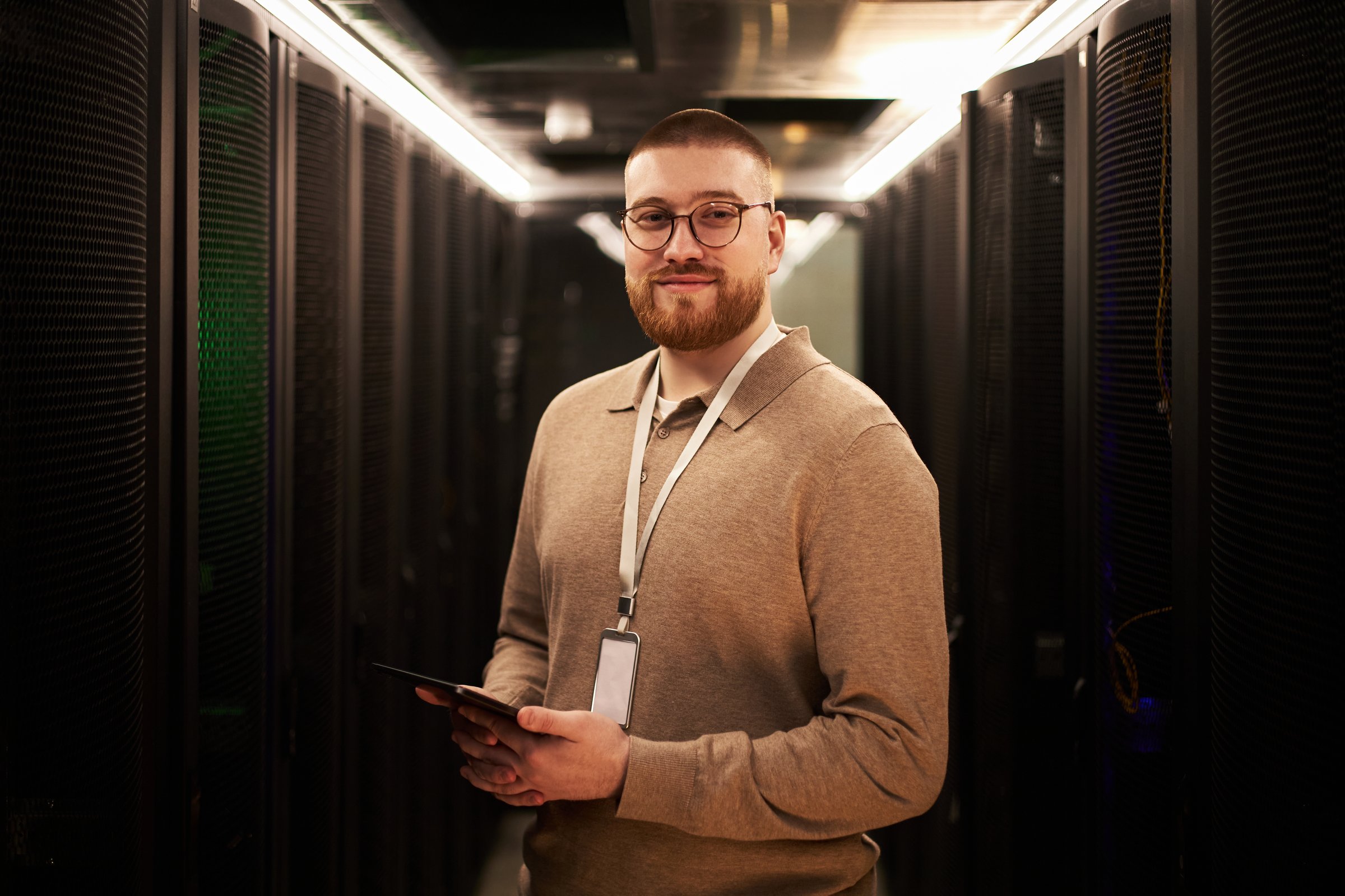 Portrait of young Caucasian man with glasses smiling and holding a tablet in a server room filled with racks of equipment and technology