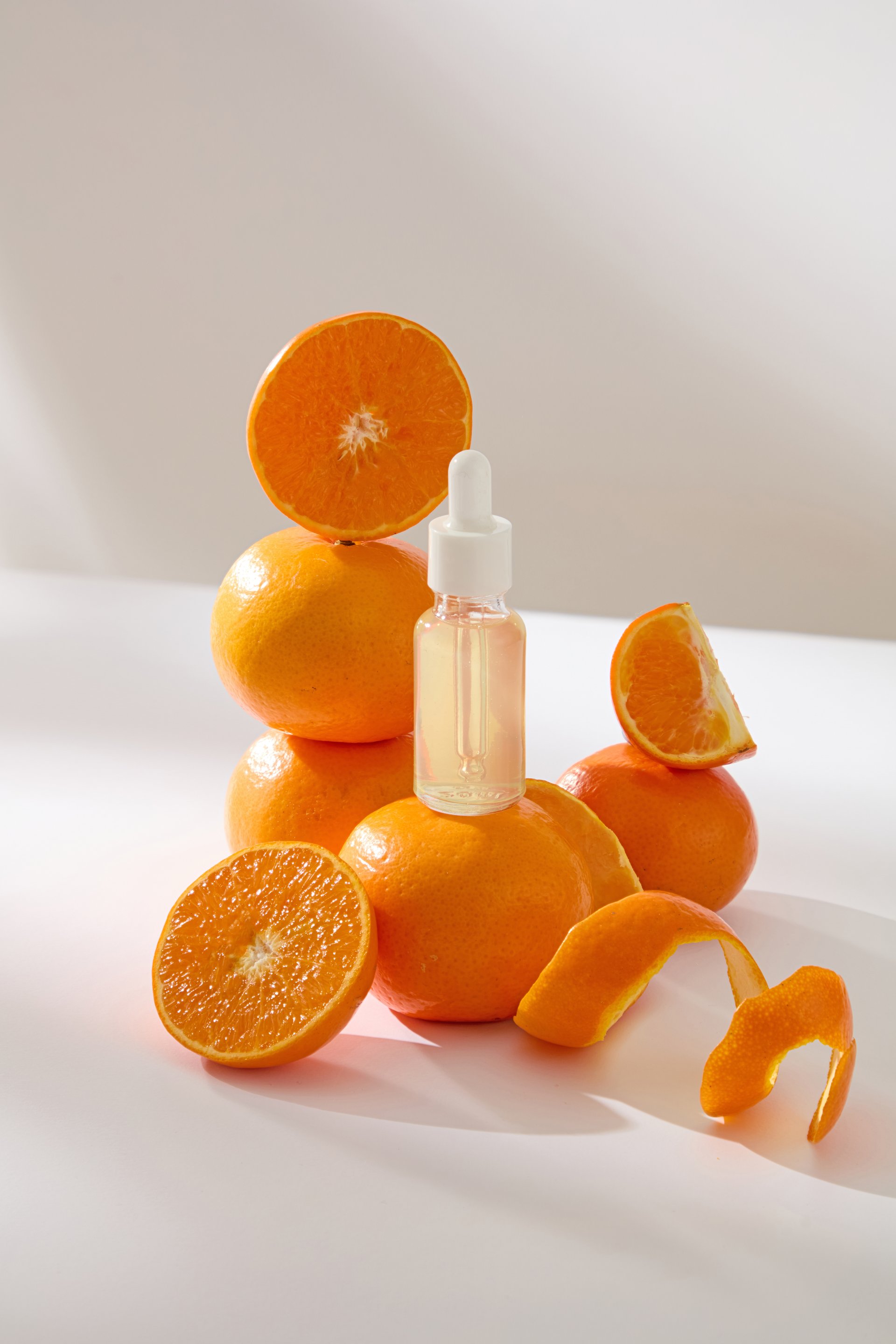 Vertical photo of transparent serum dropper bottles placed on ripe citrus. Three oranges are stacked high and others cut into different shapes. Orange peels are thinly sliced to spiraled.