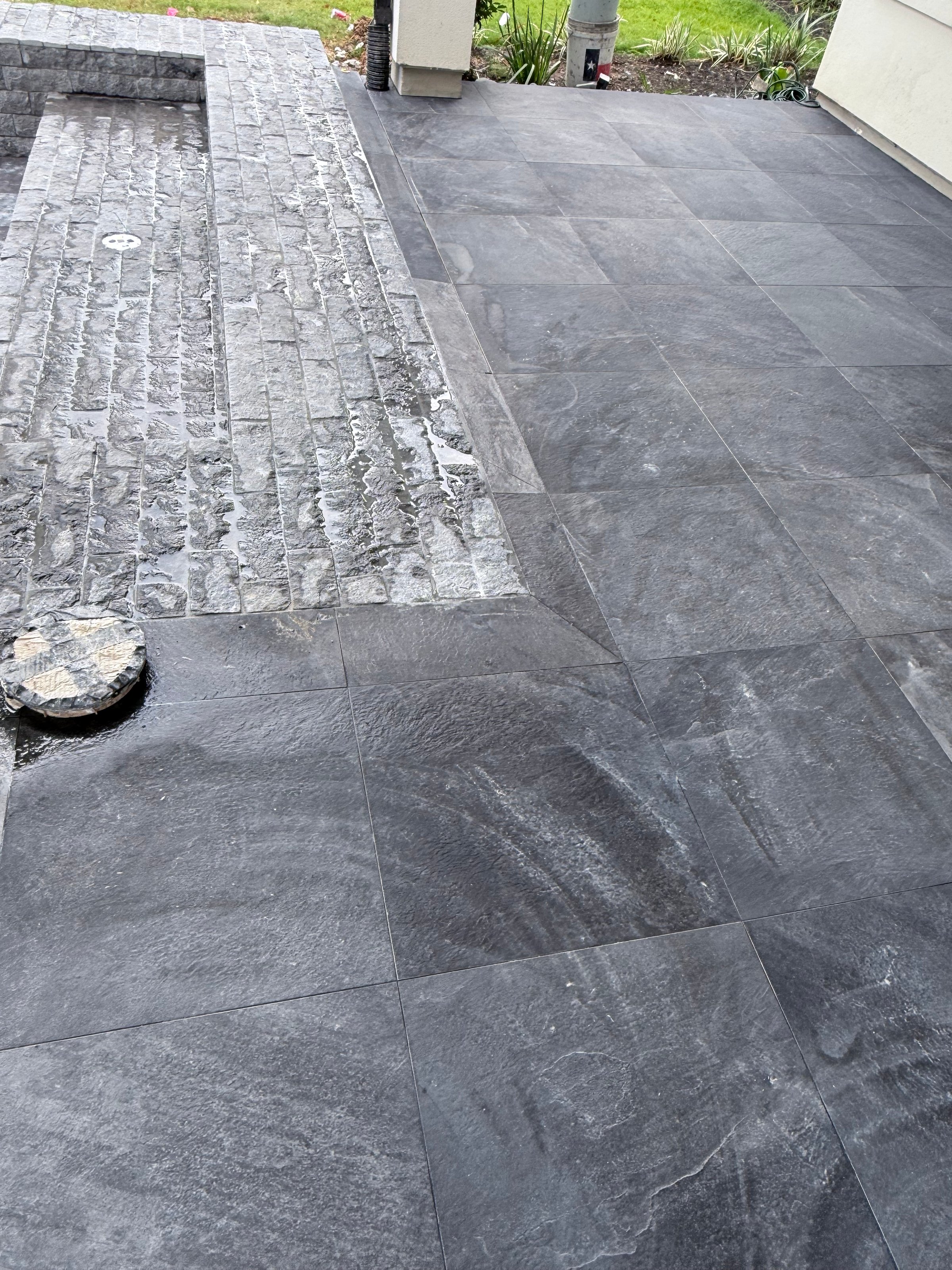 A patio with dark stone tiles, featuring a wet surface and a small circular design near the edge. Part of a building is visible.