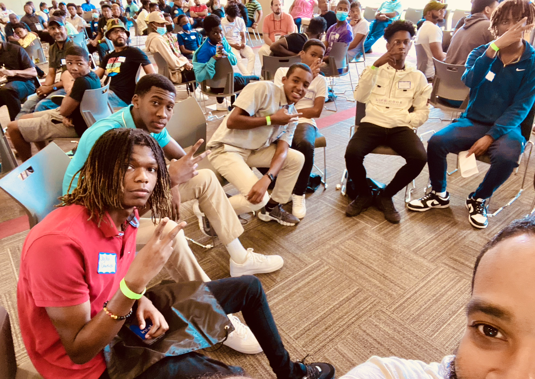 A group of young people seated in a room, some making peace signs, with others in the background attending a gathering.