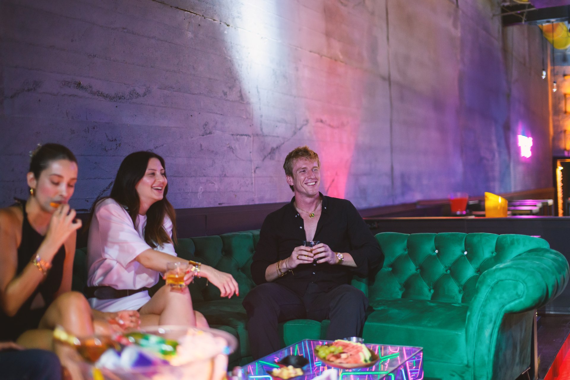 A photo of two girls and one guy, a young group lounging in a nightclub VIP area, savoring their drinks and enjoying a lively festive atmosphere.