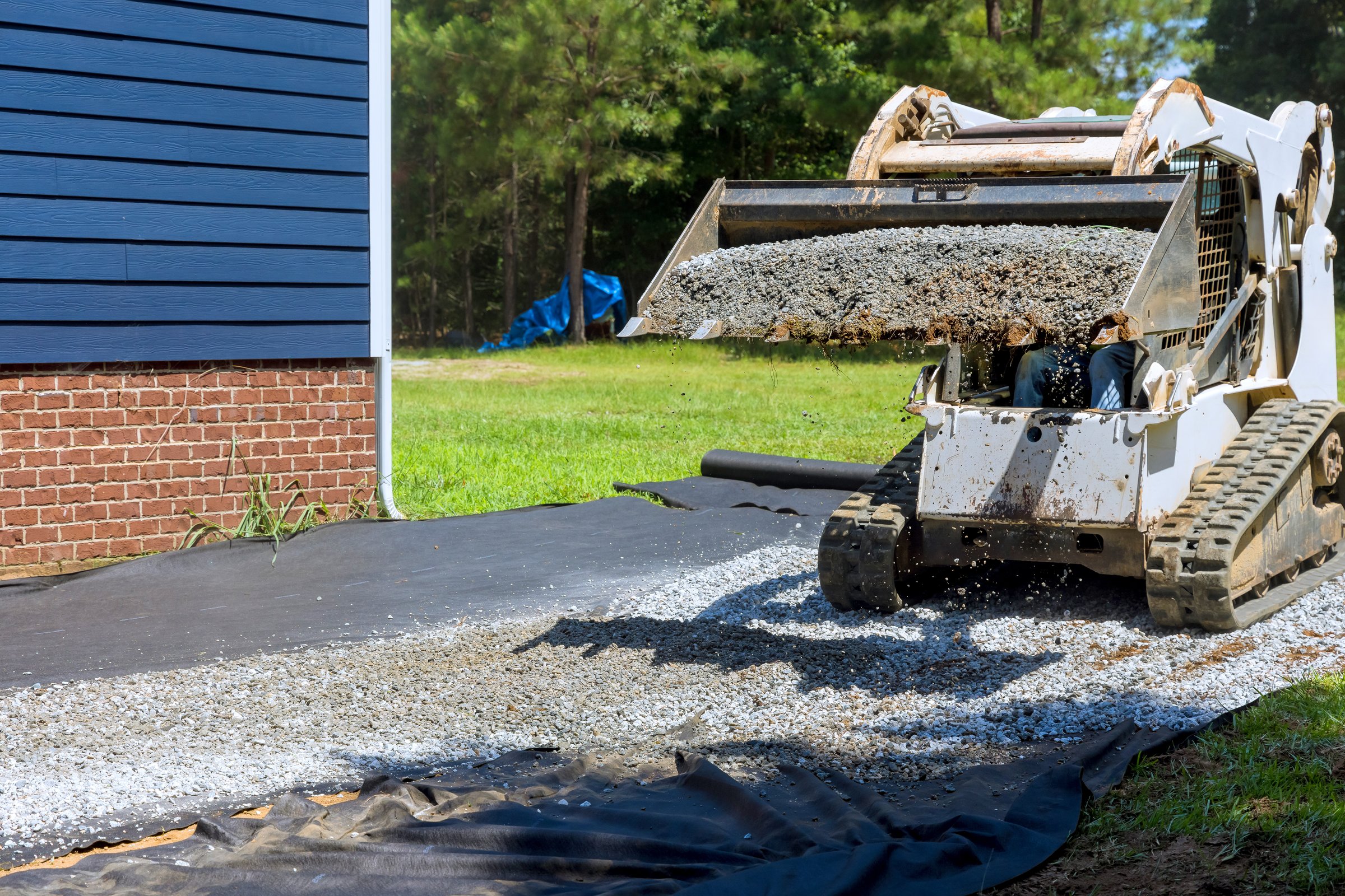 Tractor spreads gravel on black tarp beside house, preparing area for leveling landscaping during work.