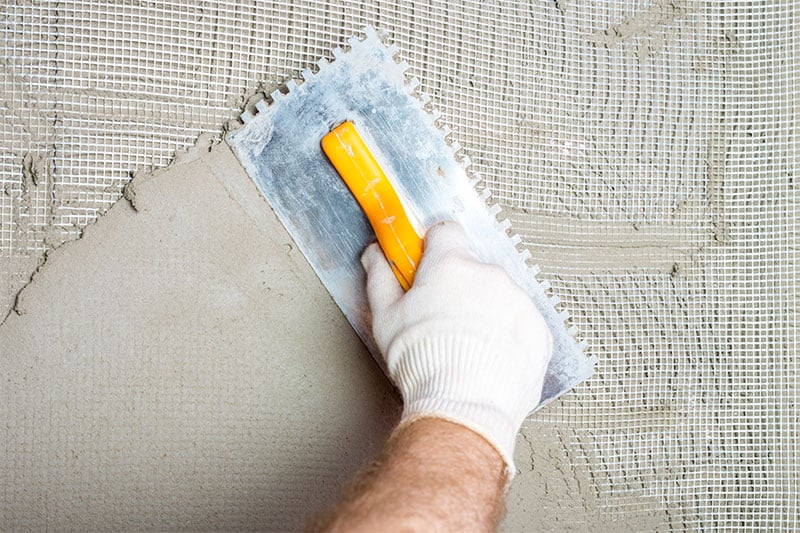 Hand in glove applying adhesive with trowel on mesh-covered surface, preparing for tile installation.