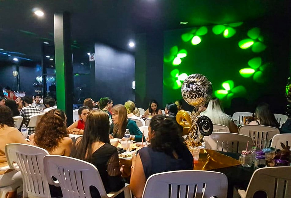 People at a party gathered around a table with balloons and festive decorations in a dimly lit room.