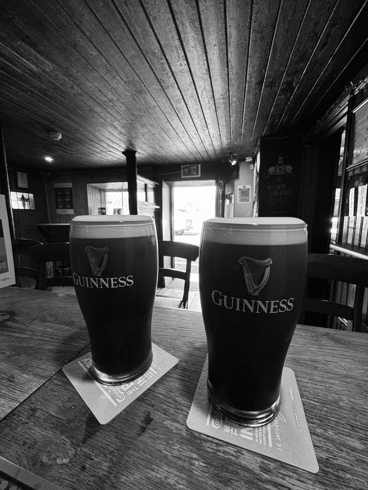 Two pints of Guinness on coasters sit on a wooden table in a dimly lit pub with a doorway visible in the background.