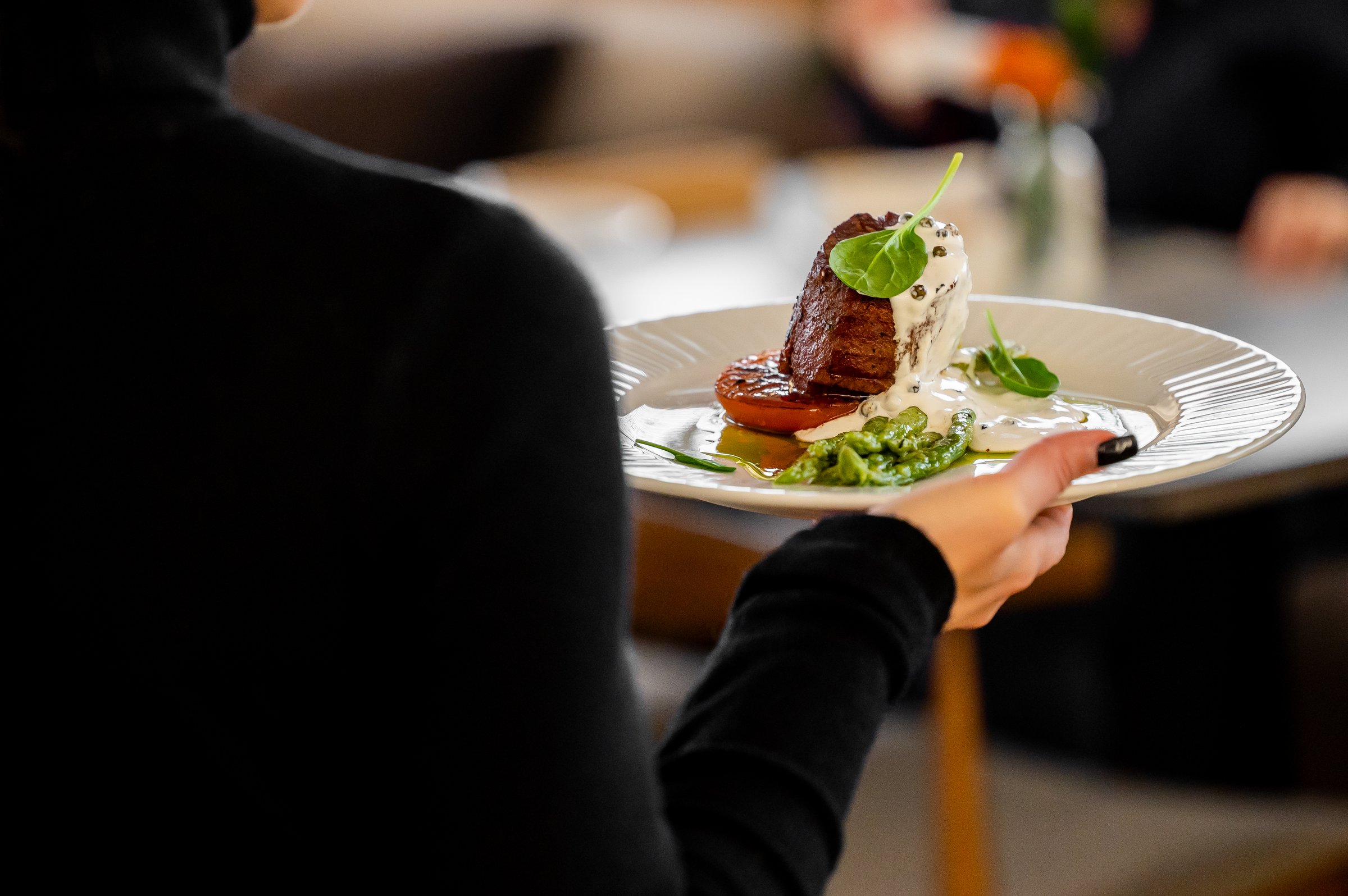 An elegantly plated gourmet dish, possibly a beef fillet with roasted vegetables and a creamy sauce, is being held by a server in a fine-dining restaurant