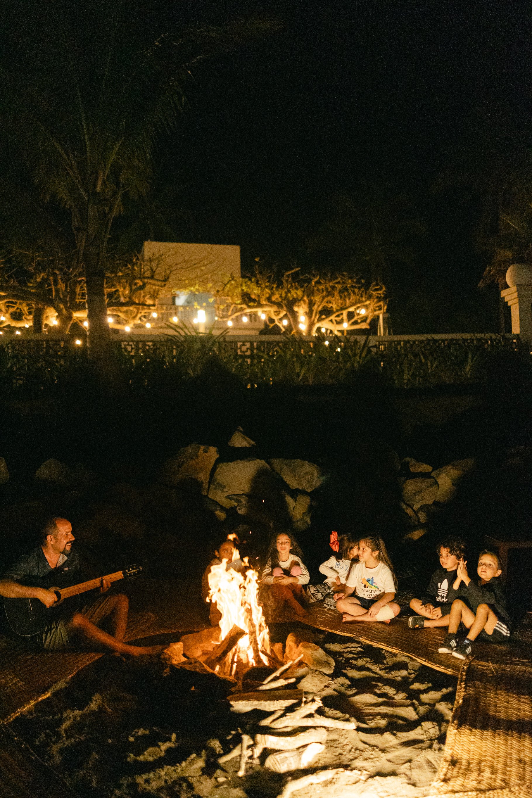 Group of people sitting around a bonfire at night, with one person playing guitar, surrounded by trees and lights.