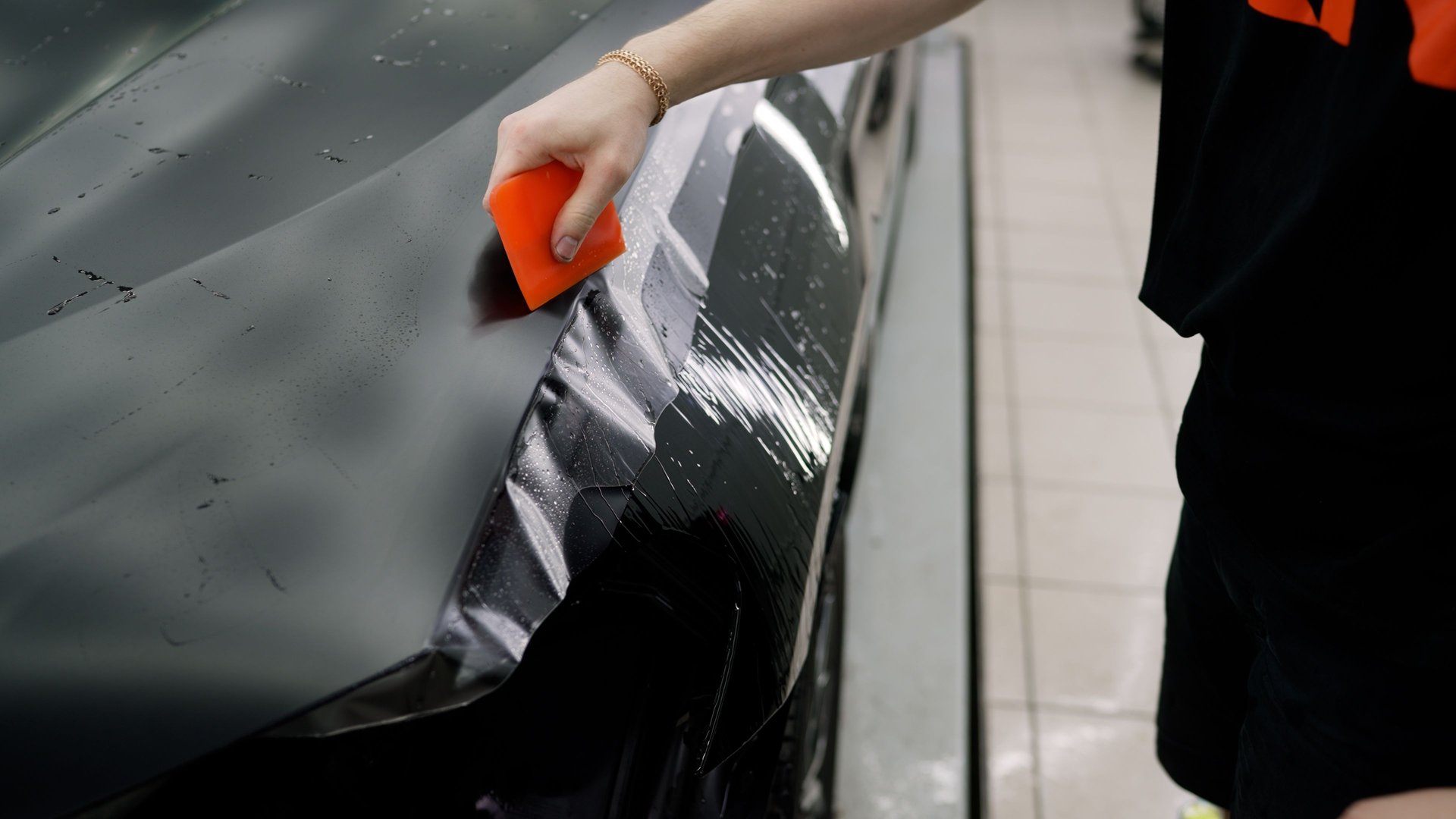 Skilled automotive professional carefully protecting car hood surface by precisely applying transparent vinyl film using specialized squeegee technique