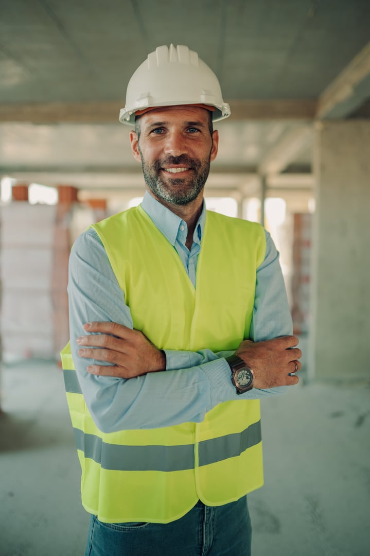 Confident engineer smiling at the camera with arms crossed, wearing a hardhat and safety vest, standing in a building under construction
