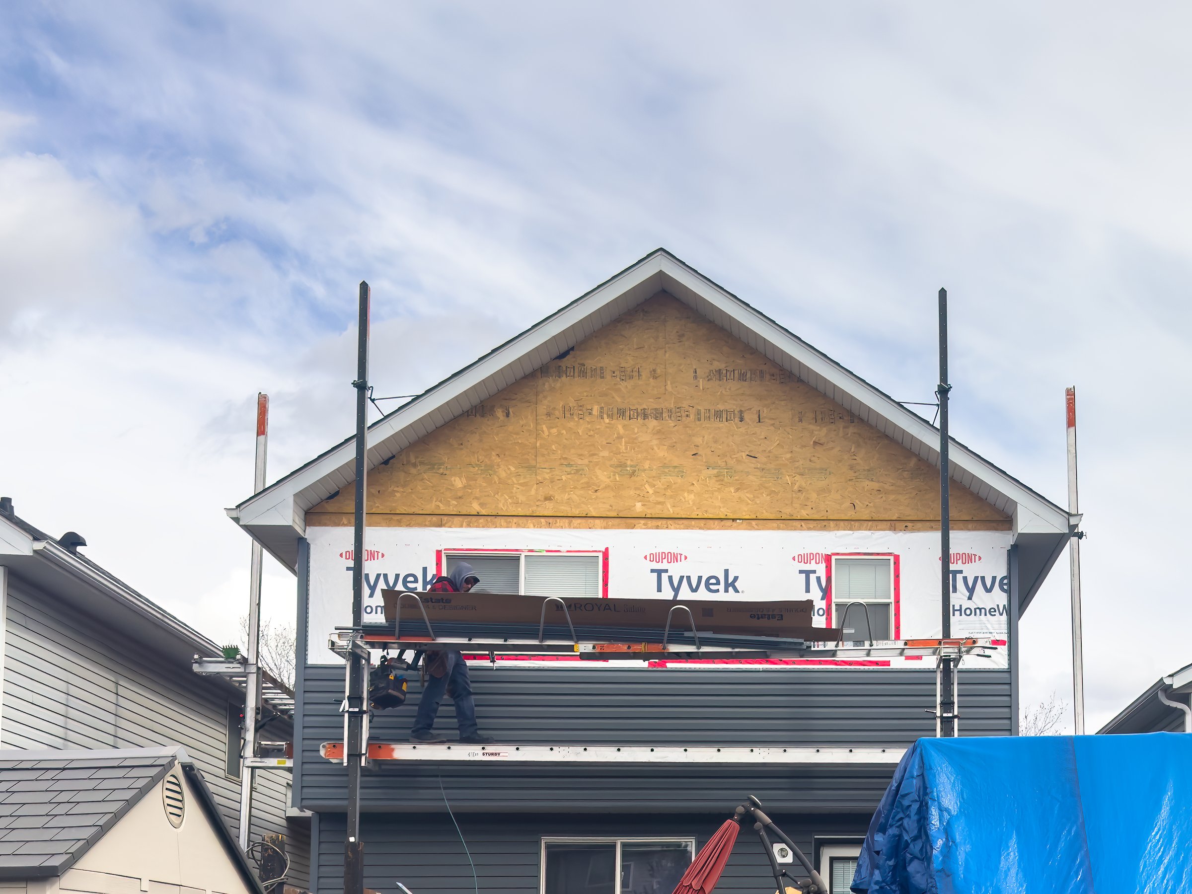 Calgary, Alberta, Canada. Apr 10, 2025. A house under construction shows gray siding being installed over white Tyvek wrap. Workers are on scaffolding against a cloudy sky.