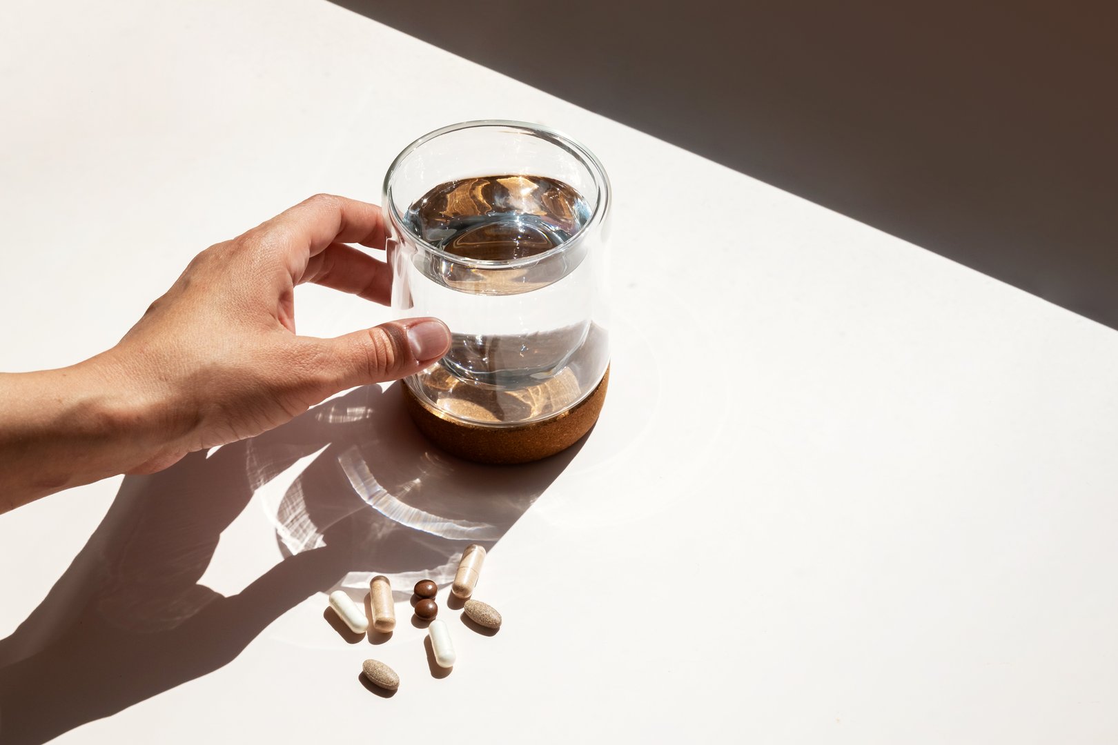 Person Reaching for Water Glass next to Scattered Vitamin Pills. Daily Supplement Intake for Health and Wellness.