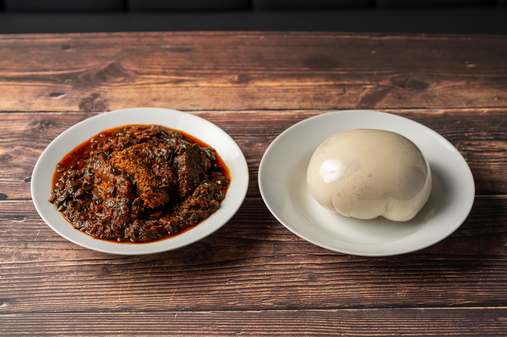 A plate of traditional African vegetable soup with meat served alongside a plate of fufu on a wooden table.