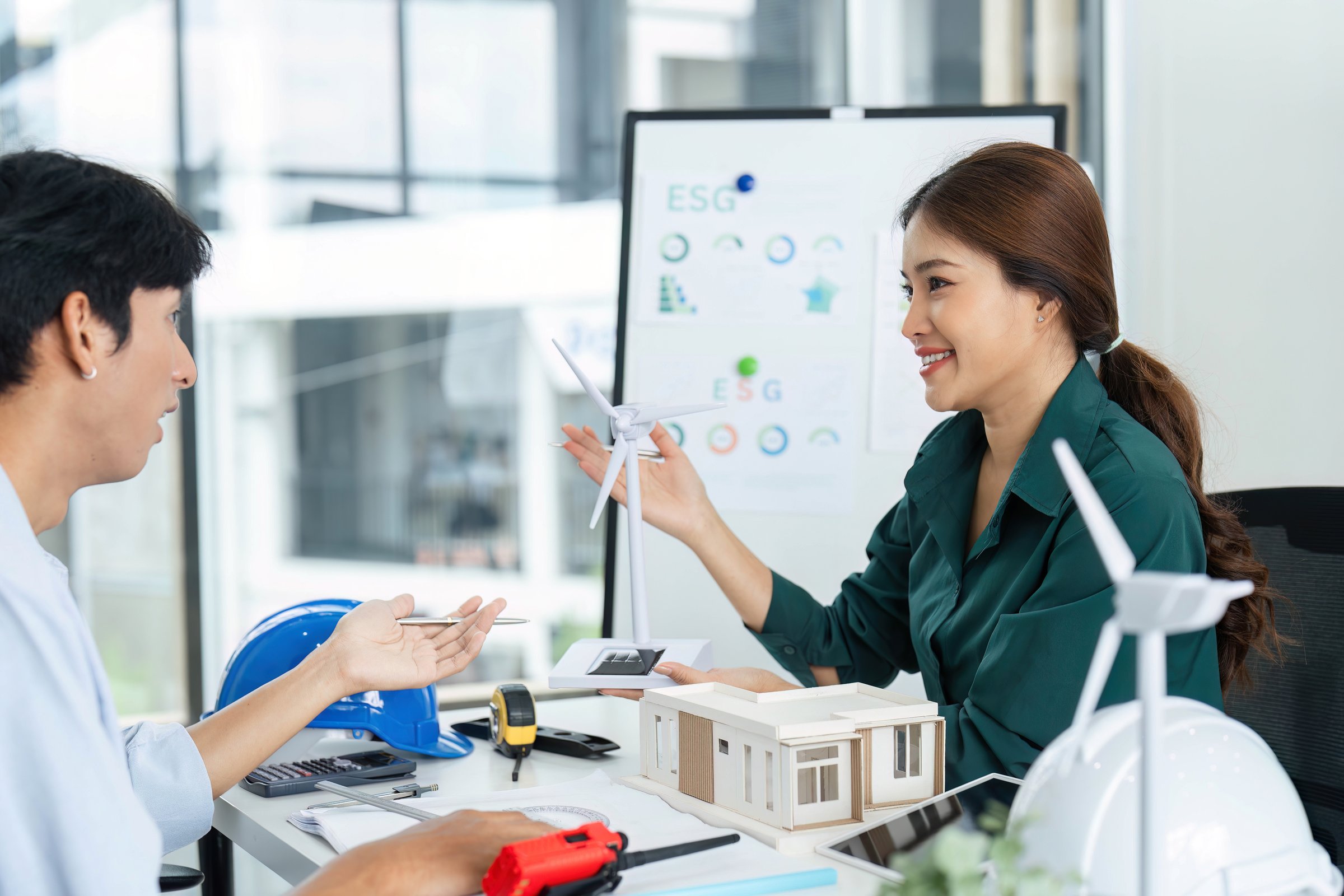 A woman presents a model while discussing ESG strategies with a colleague, emphasizing renewable energy and sustainable design in a modern workspace.
