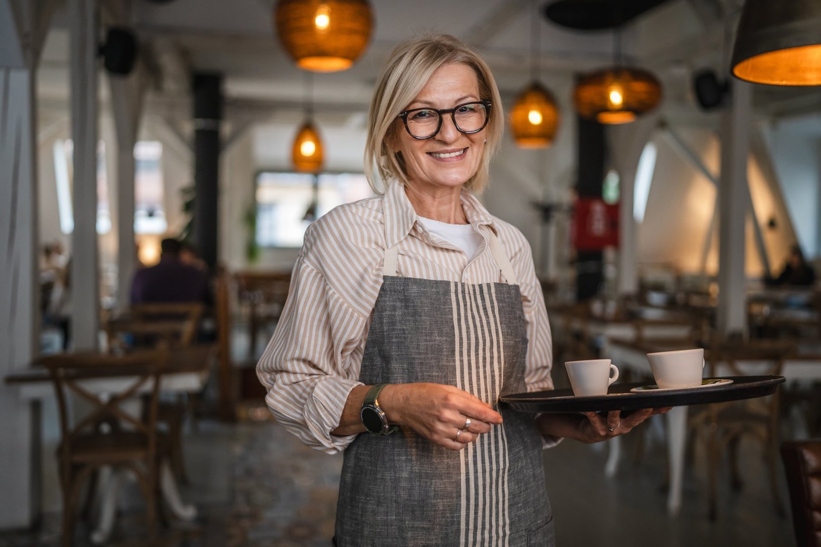 Nice mature woman waitress in apron offer cup of delicious tasty coffee, stand and smile, friendly staff of restaurant