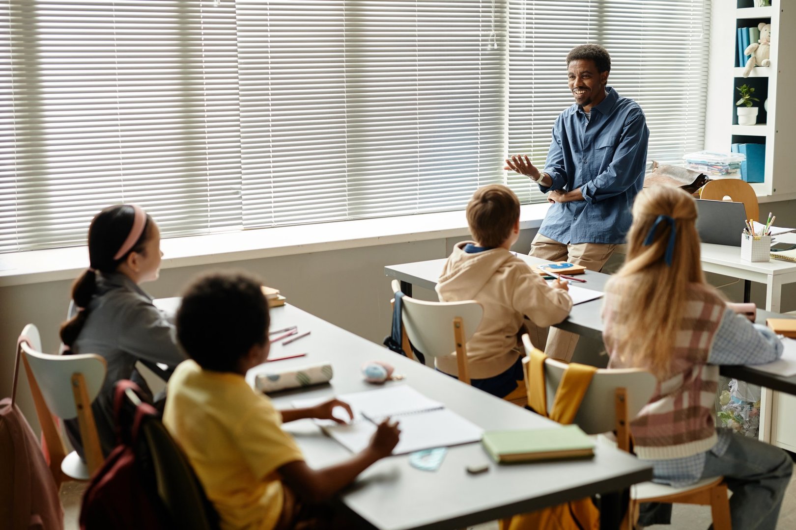 Wide angle shot of smiling male African American teacher talking to children in classroom with window blinds while explaining new topic at primary school, copy space