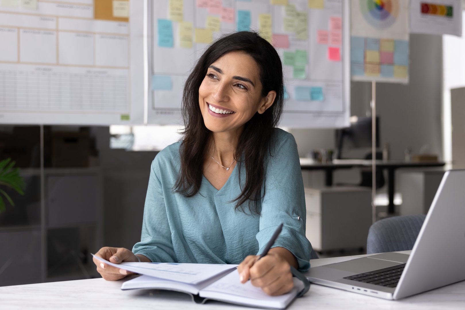 Positive young Latin manager woman working in office, analyzing paper report, writing in notebook, sitting at laptop, looking away, thinking on work schedule, planning tasks