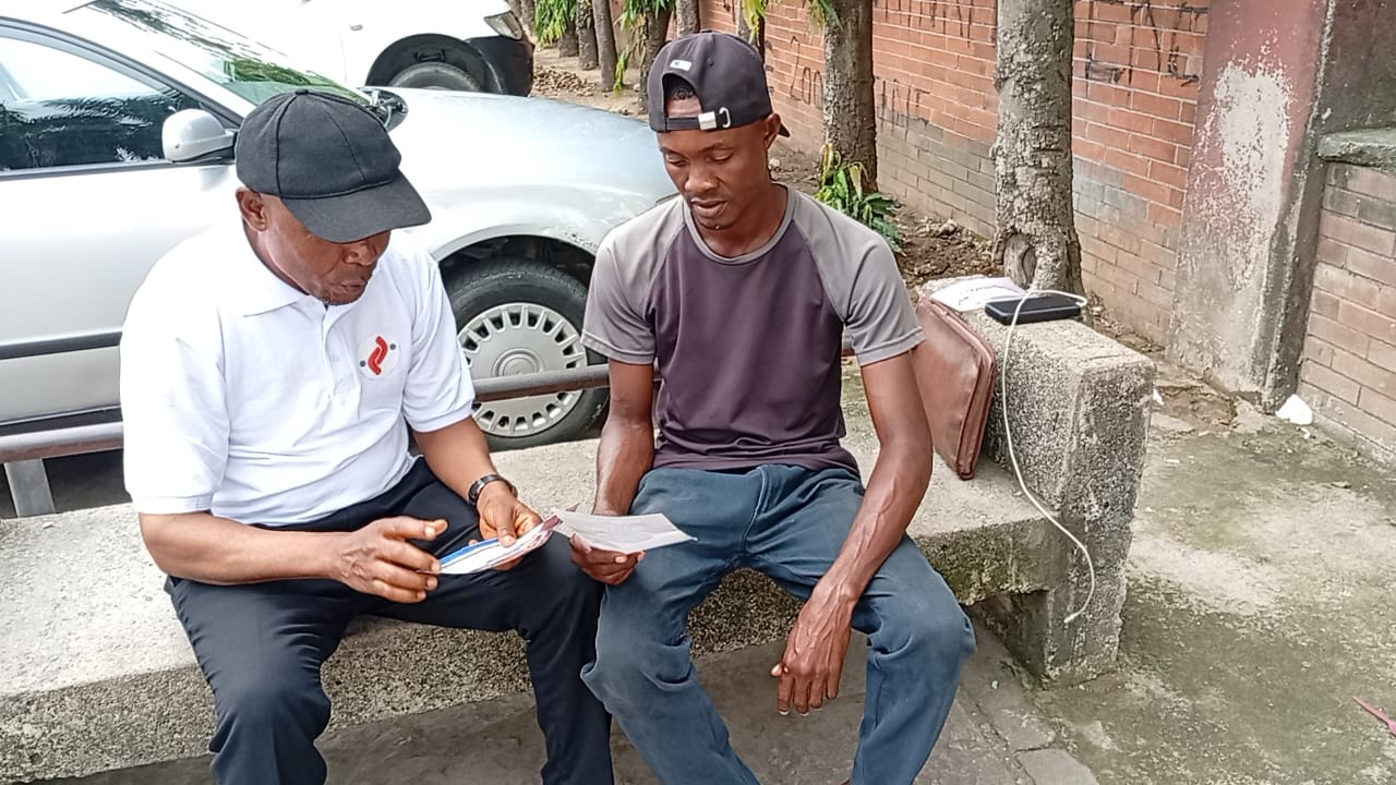 Two men sitting on a stone bench, one holding papers, engaged in a conversation near parked cars and a brick wall.
