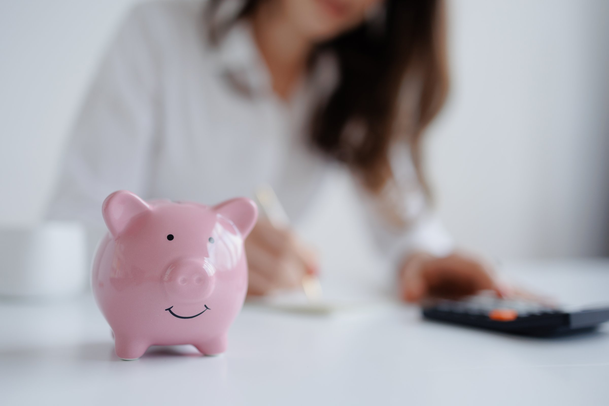 A person works at a desk, writing notes and using a calculator, while a pink piggy bank sits prominently in the foreground, symbolizing financial planning and saving.