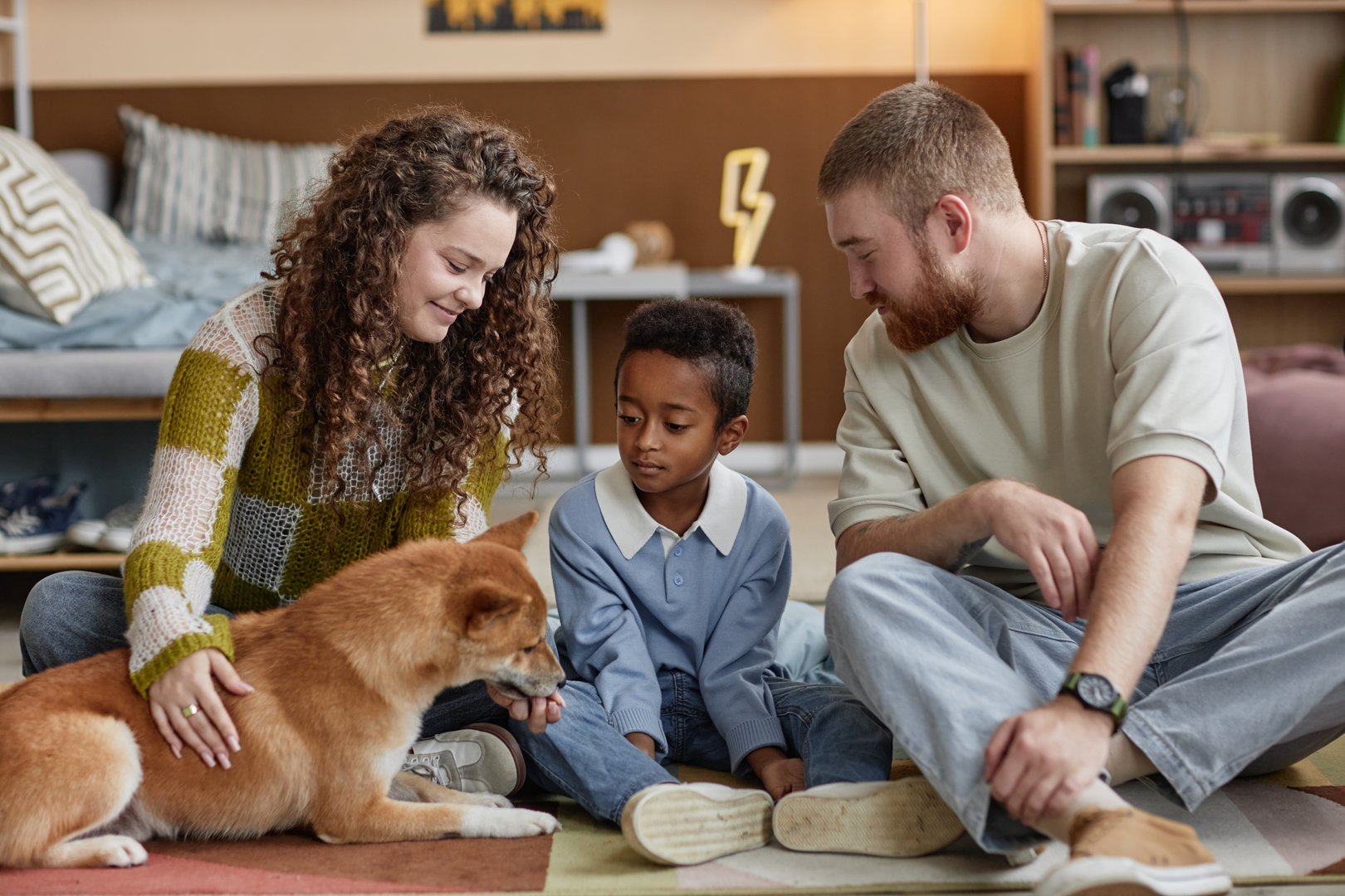 Little Black boy sitting with smiling adoptive parents while playing with Shiba Inu dog on floor in kids room, happy family concept