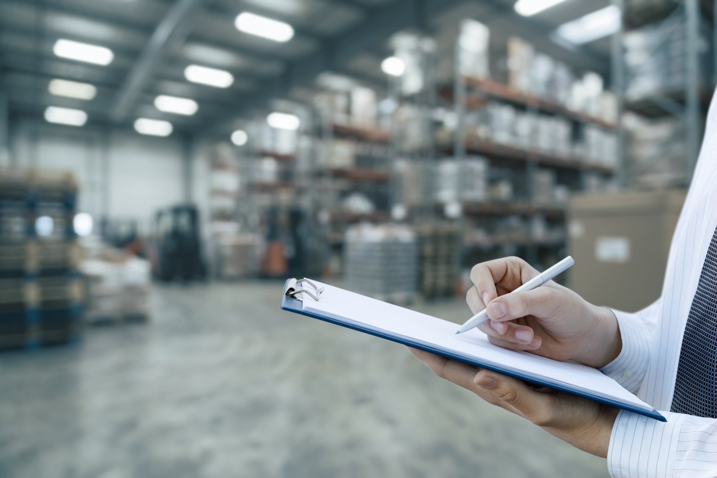 Warehouse task setup. A person writes notes on a clipboard in a bustling warehouse filled with goods.