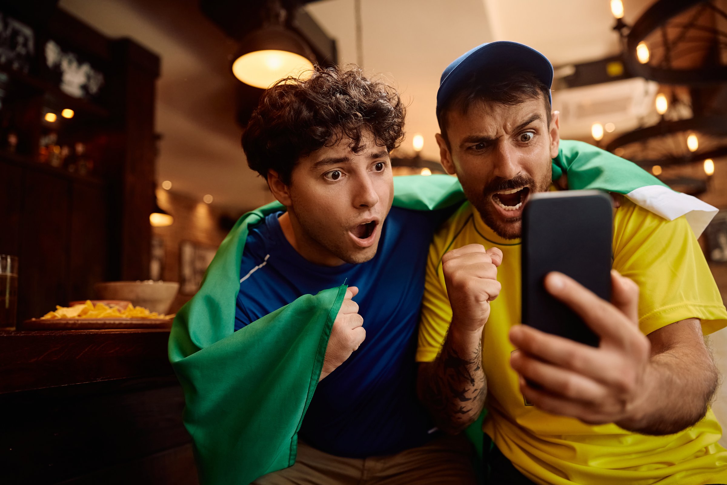 Young men cheering while watching sports match on smart phone in a pub.