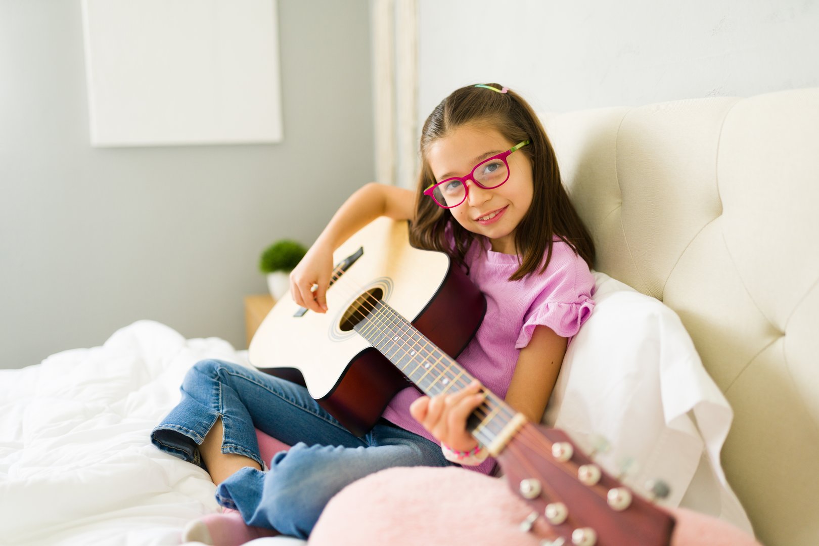 Happy little girl learning to play acoustic guitar sitting on bed in bedroom, practicing new song and enjoying music
