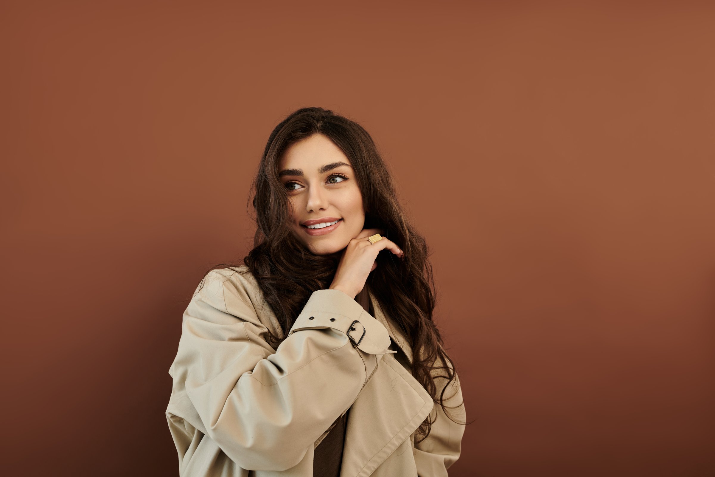 Dressed in stylish autumn attire, a beautiful young woman smiles gracefully while posing in a studio with earthy tones.