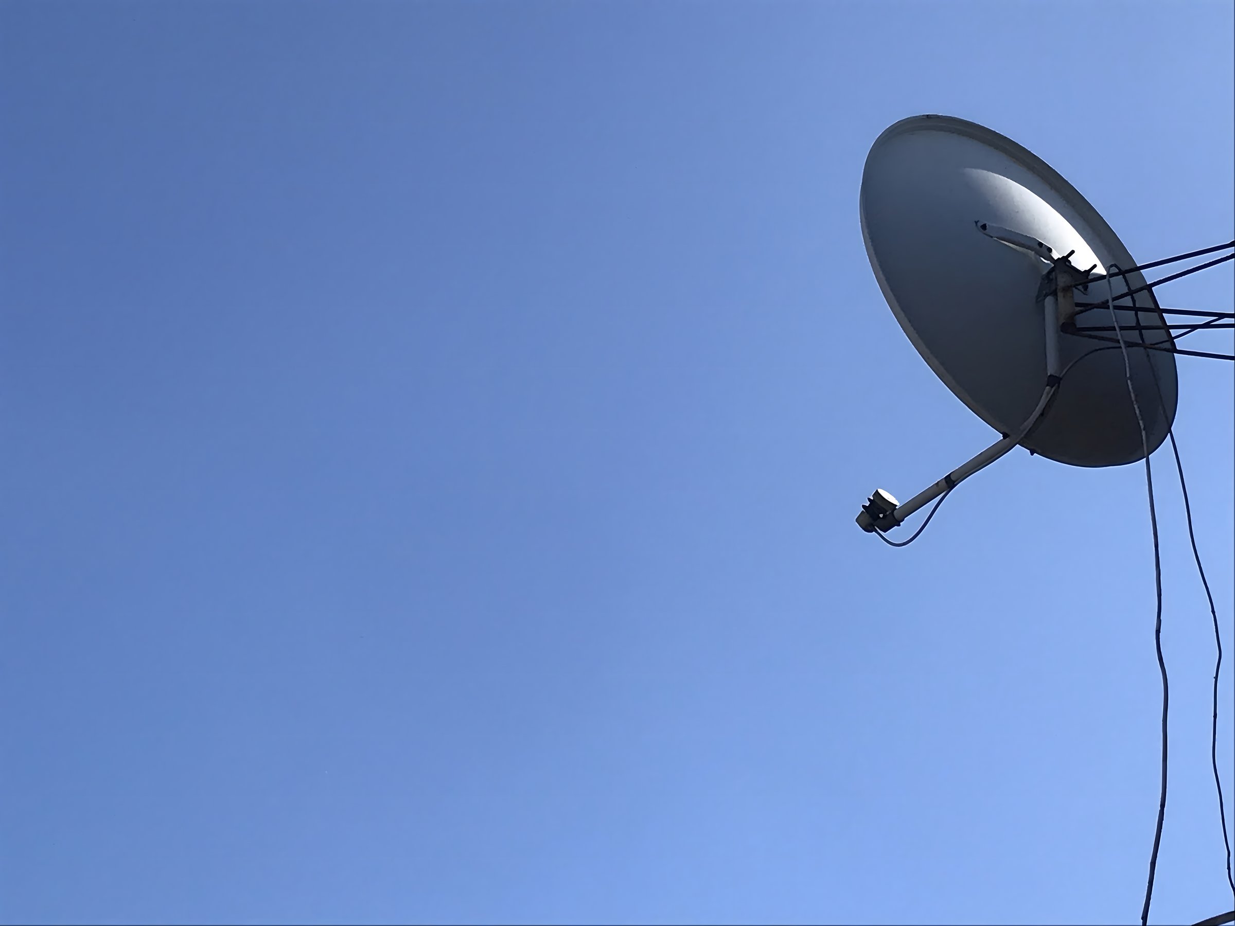 A satellite dish is mounted on a pole, capturing signals against a backdrop of a clear blue sky. The image is taken during midday in an urban environment.