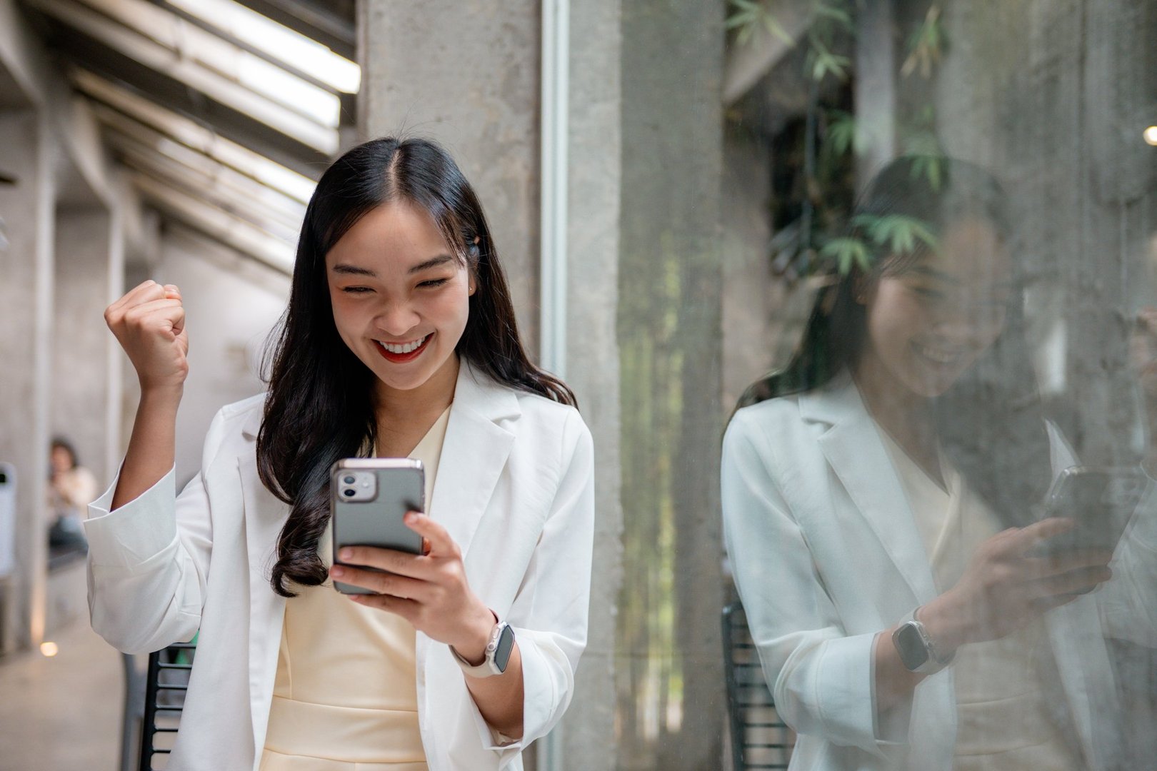 Young asian businesswoman feeling happy and raising her fist while looking at her smartphone, celebrating a successful business achievement in a modern office environment
