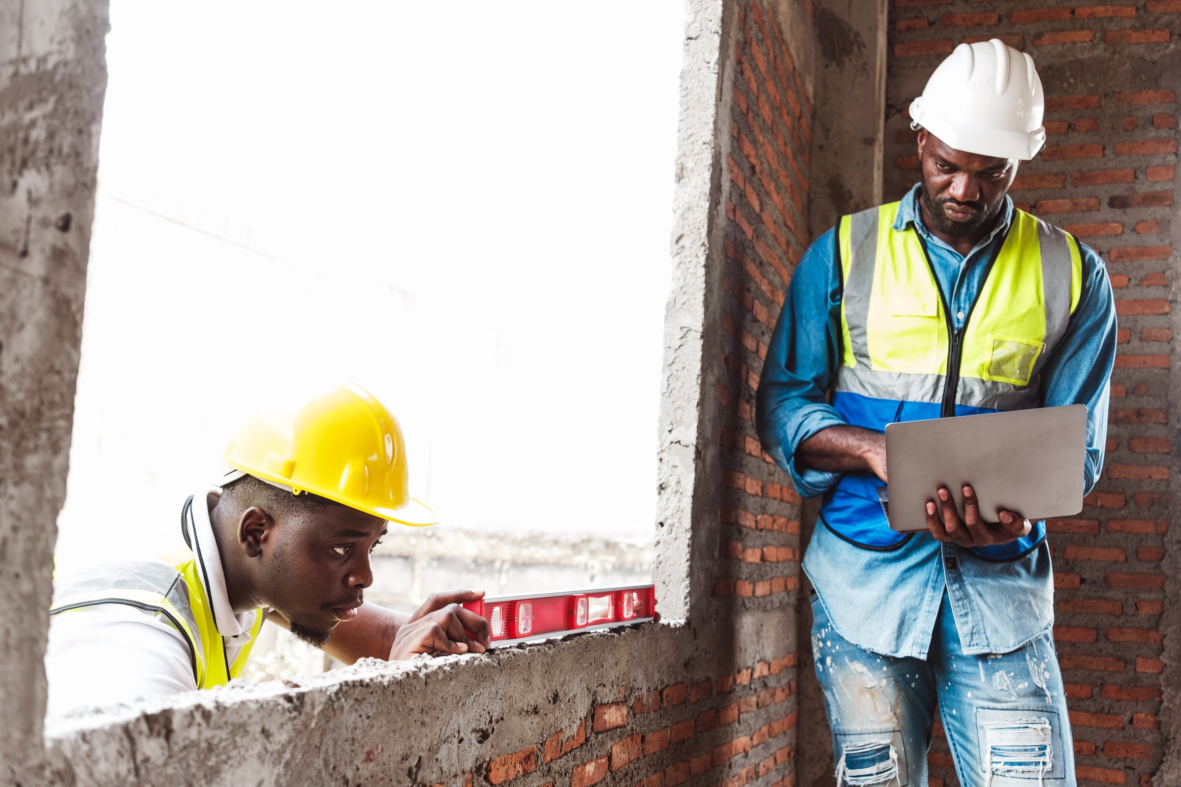 Two Black construction workers collaborate at a building siteone using a spirit level, the other reviewing blueprints on a laptop. Image highlights teamwork, skill, and modern construction workflow.
