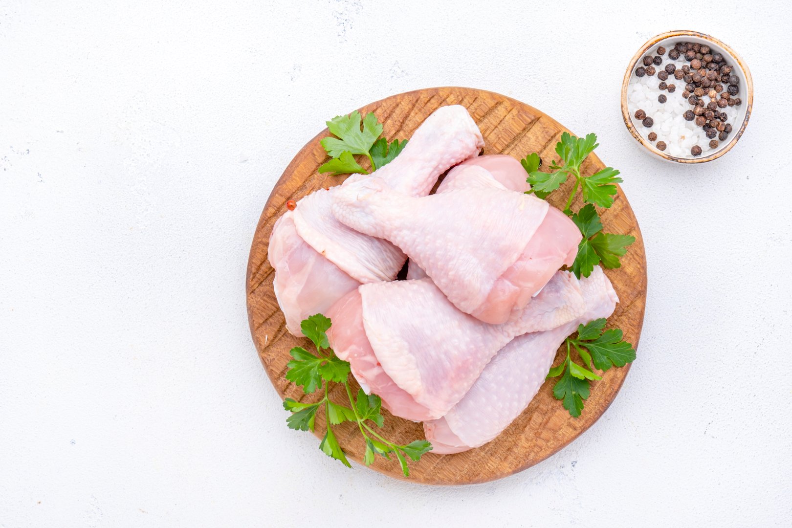 Raw chicken legs on round wooden board with pepper, salt and parsley. White background, top view