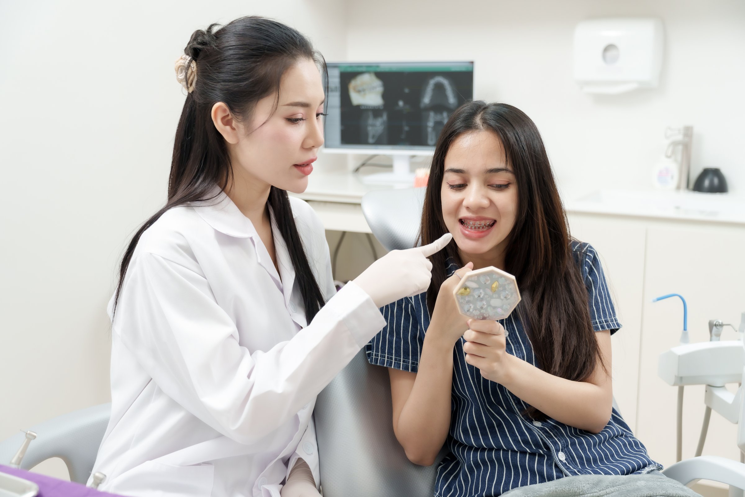 Multiracial female patient with braces holding tooth model while asian female dentist explains correct oral hygiene techniques during dental consultation in modern clinic with X-ray screen and chair