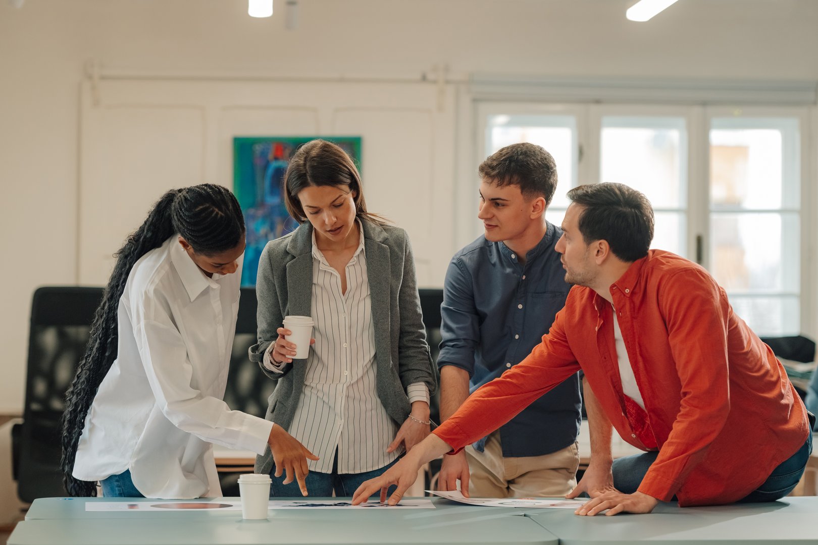 Four young professionals, two women and two men, are examining marketing charts and discussing strategy in a contemporary office setting, with one woman holding a coffee cup