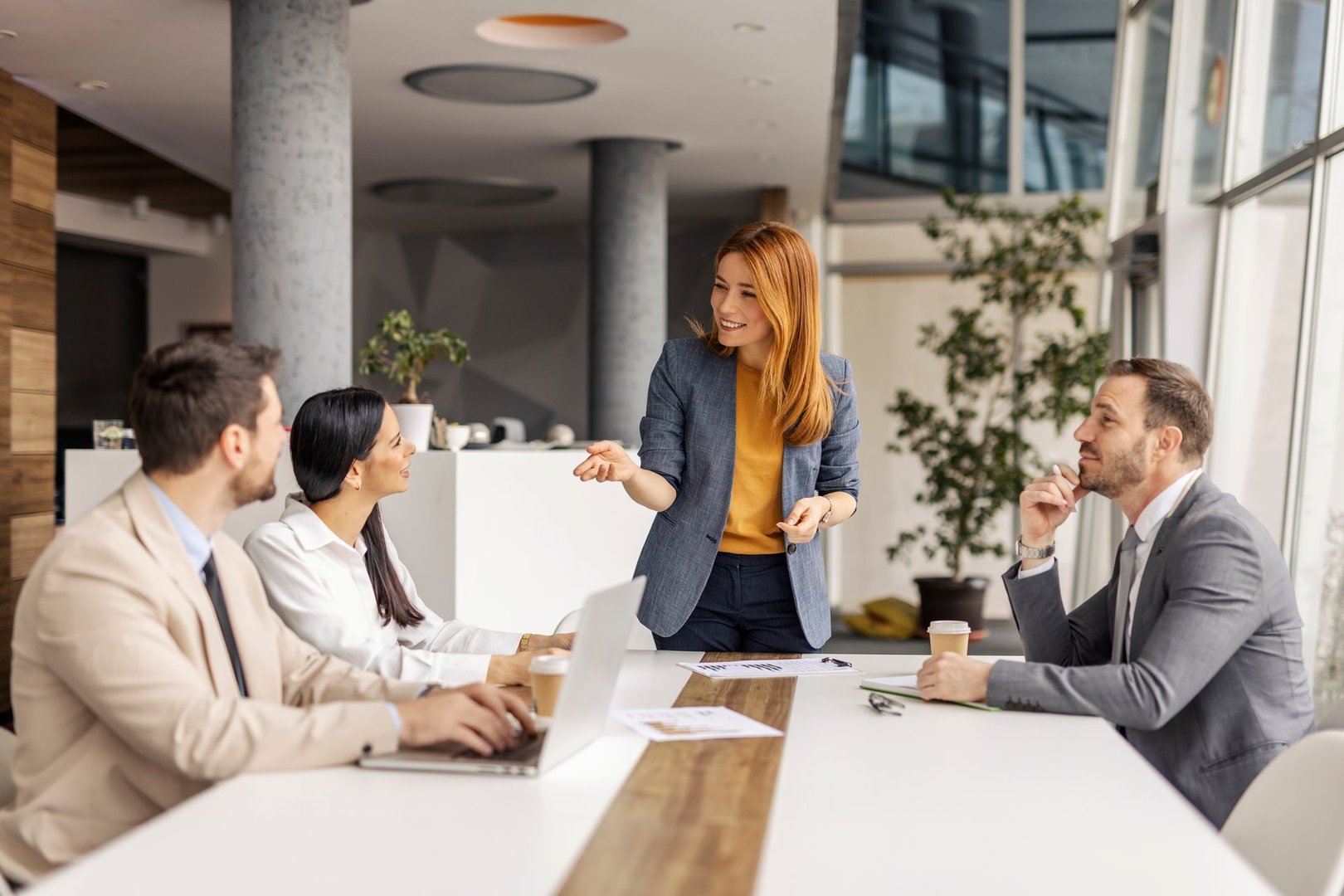 Smiling female manager standing at boardroom at corporate office with her team and discussing project with her colleagues on a meeting.