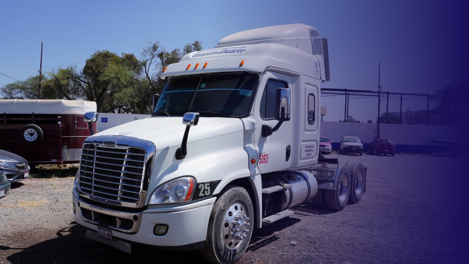 White semi-truck parked in a gravel lot, with a horse trailer and other vehicles in the background.