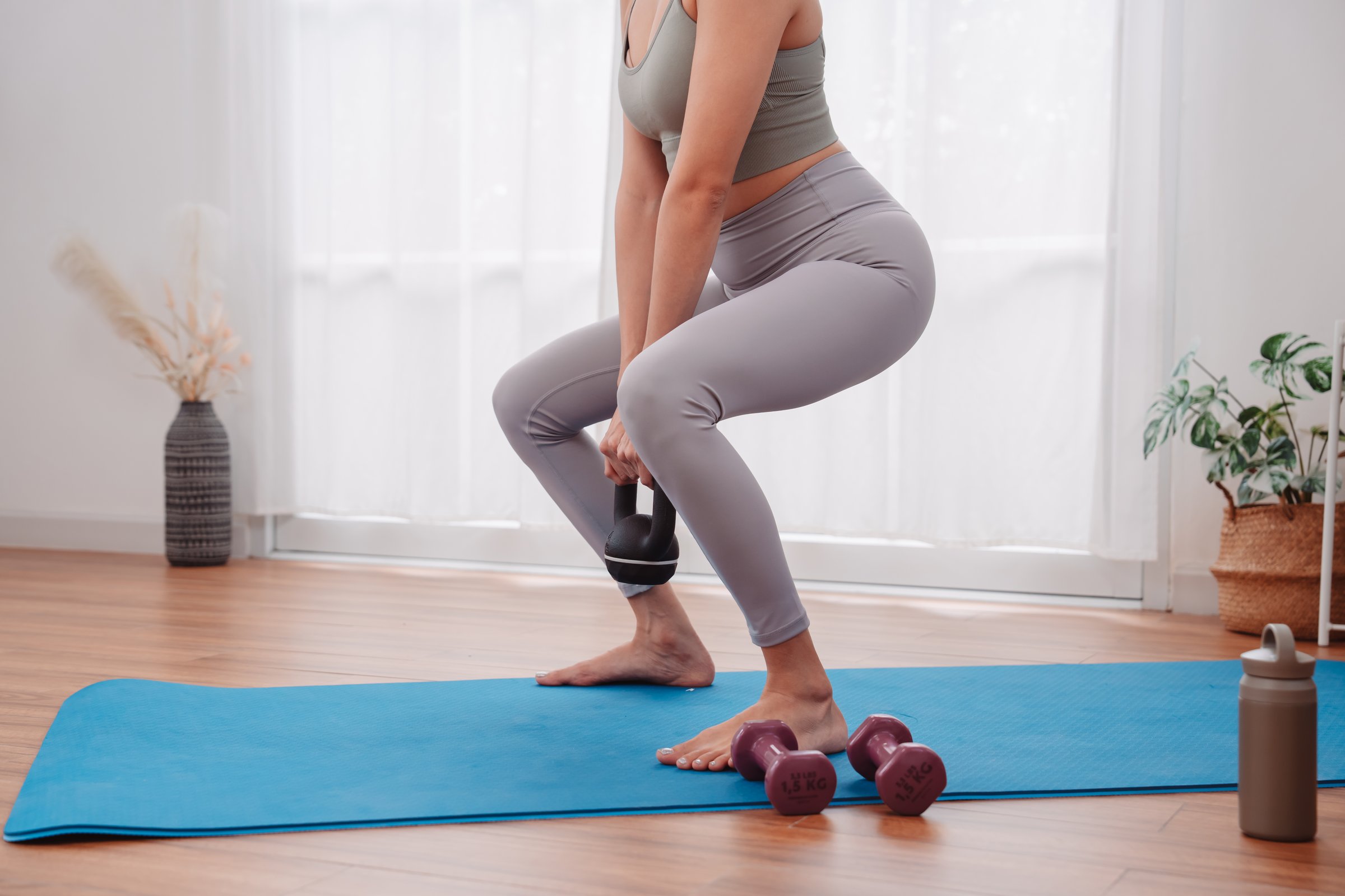 Squat exercise with kettlebell by Asian woman at home on yoga mat, strength training, lower body workout, and female fitness routine with minimal gym equipment.