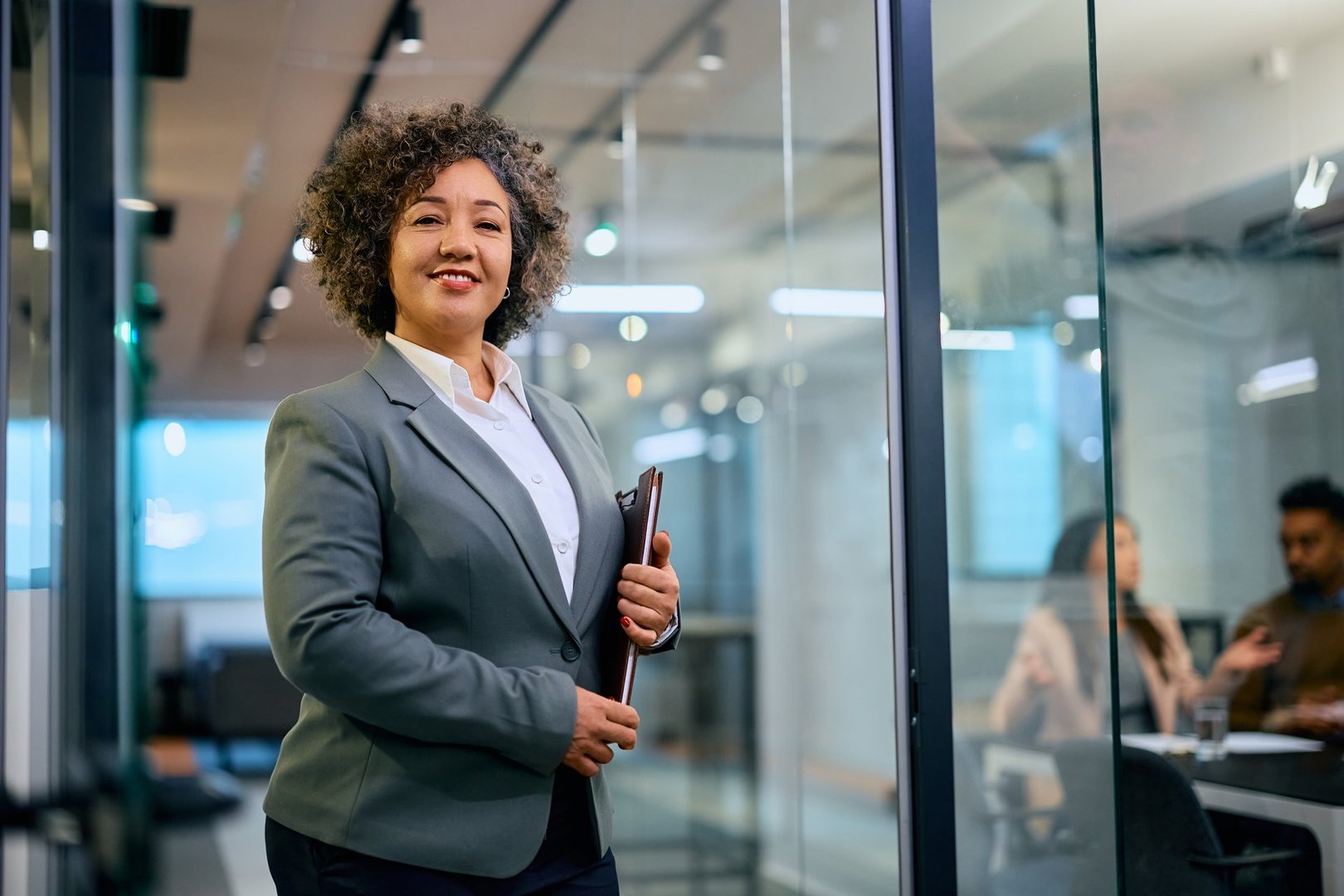 Happy financial consultant at work looking at camera. Her clients are in the office in the background.