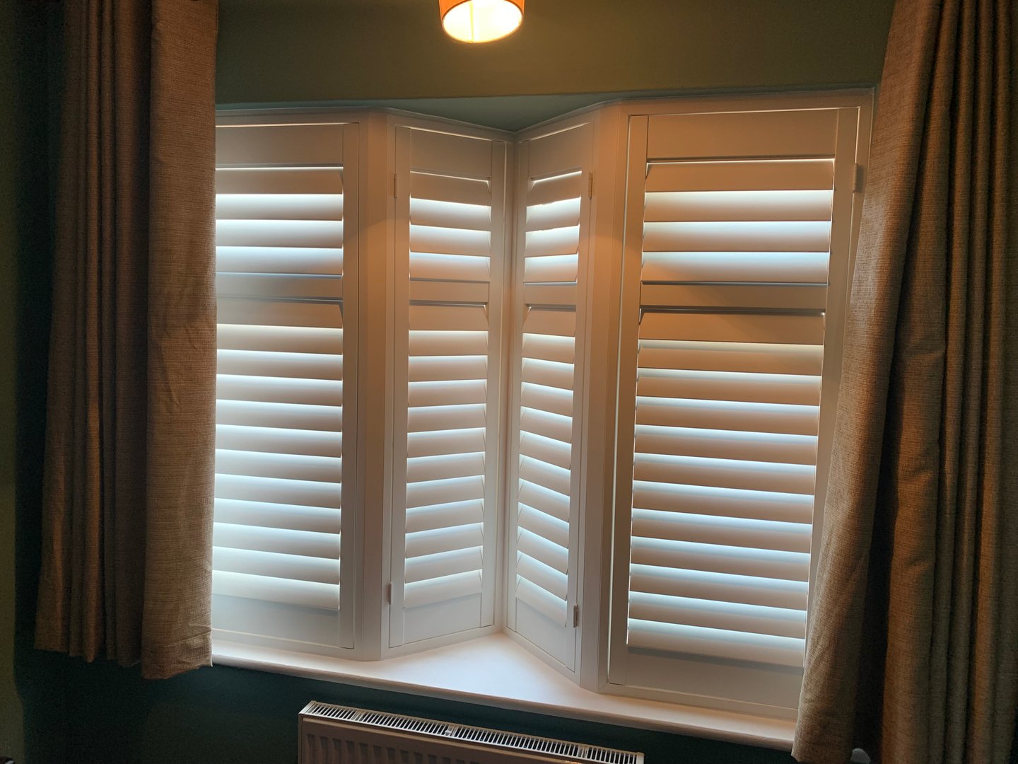 A bay window with white plantation shutters partially open, surrounded by dark curtains and above a radiator.