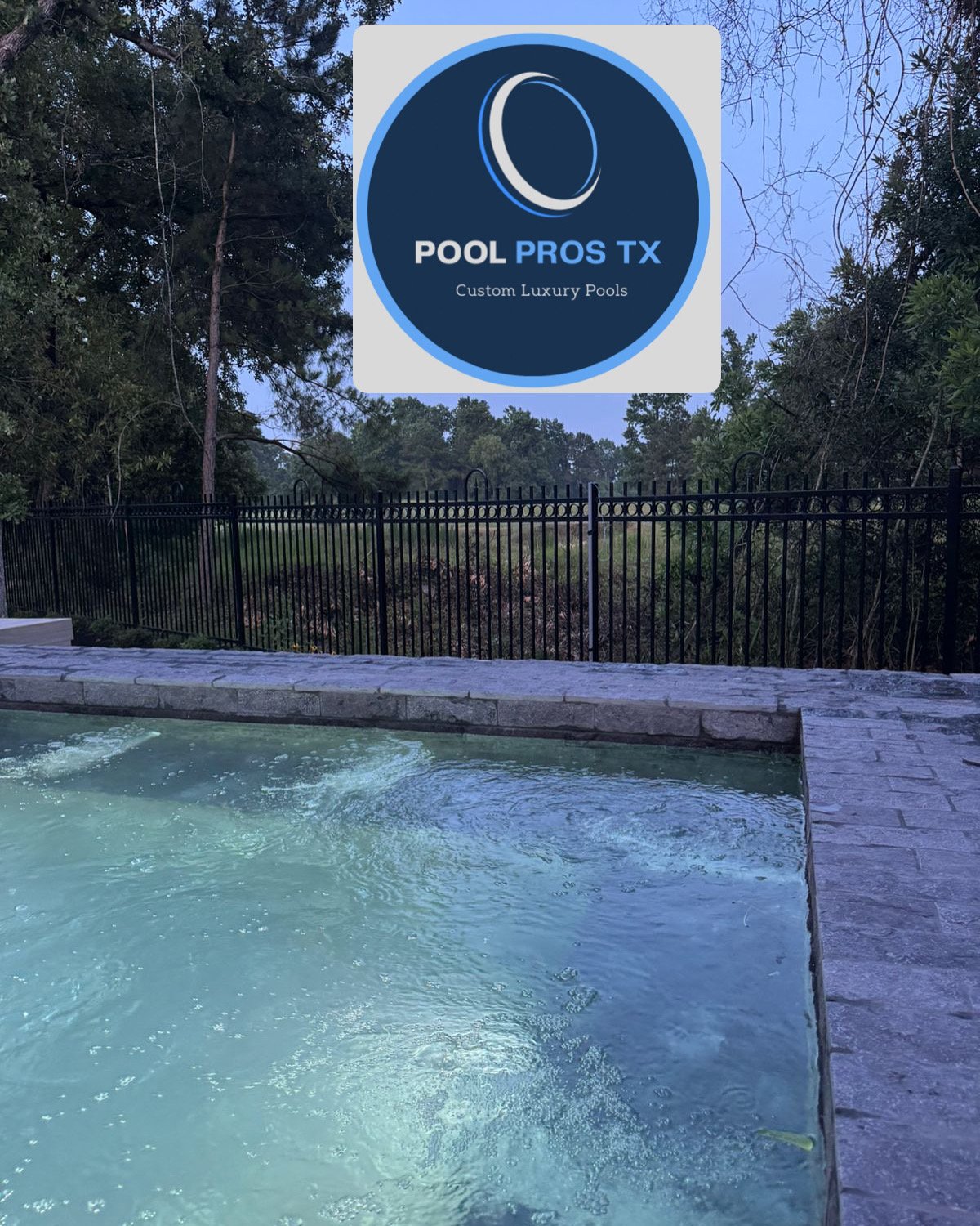 Swimming pool with stone edge at dusk, surrounded by a black fence, with a logo Pool Pros TX: Custom Luxury Pools above.