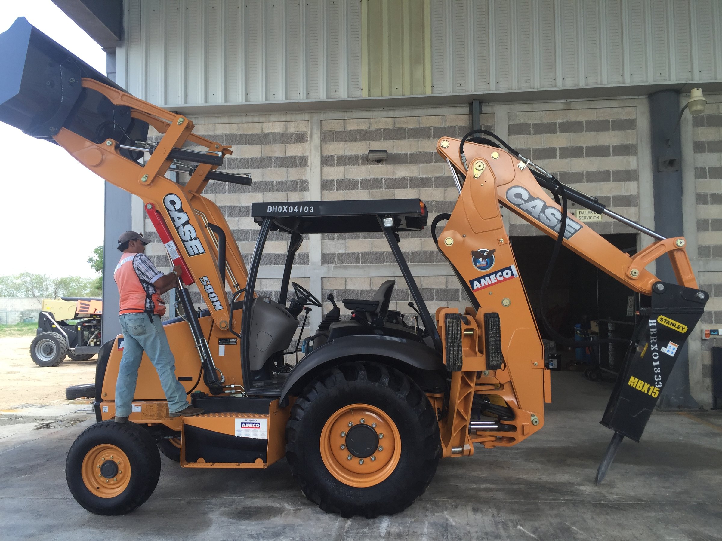 Man standing beside an orange CASE backhoe loader with machinery attachment, parked in an industrial area.
