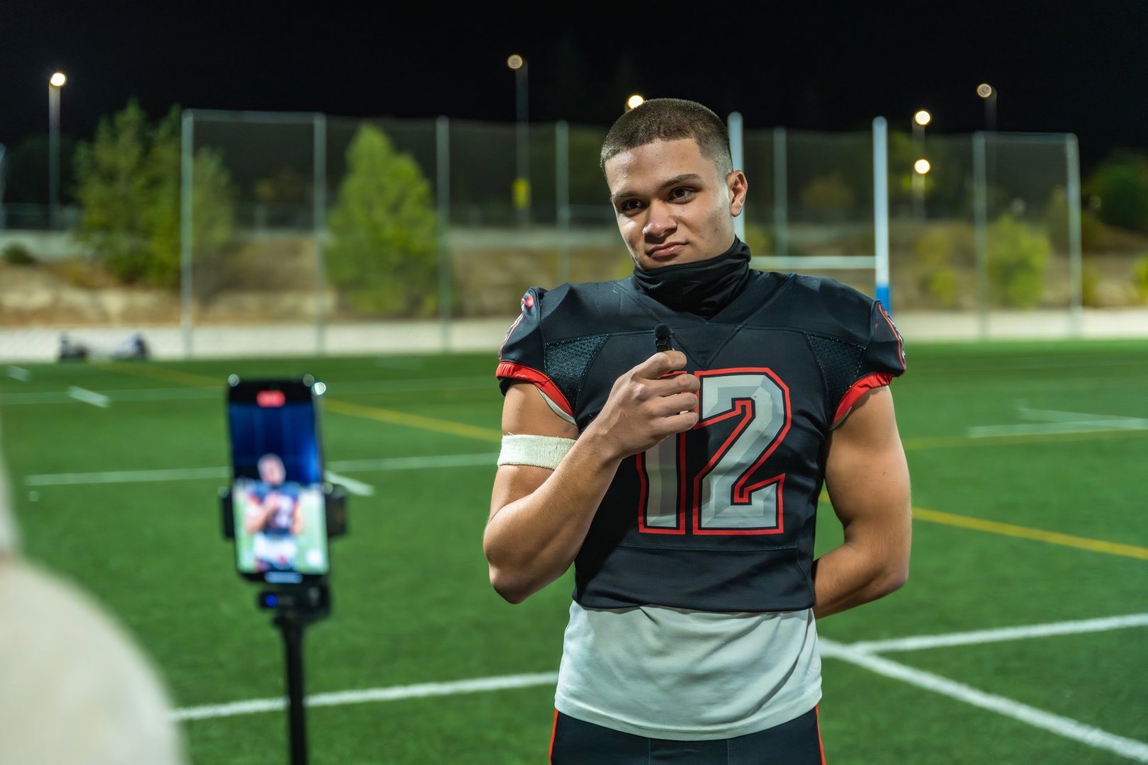 Young male football player in uniform stands on a lit stadium field at night holding a microphone while doing a live-stream interview on a smartphone, looking at the camera for broadcast content
