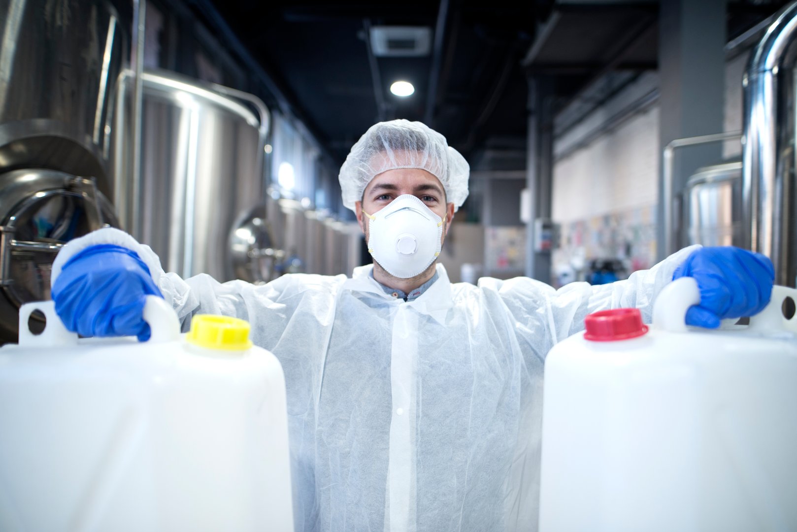 Industrial worker with protective mask and white uniform holding plastic cans for chemical industry.